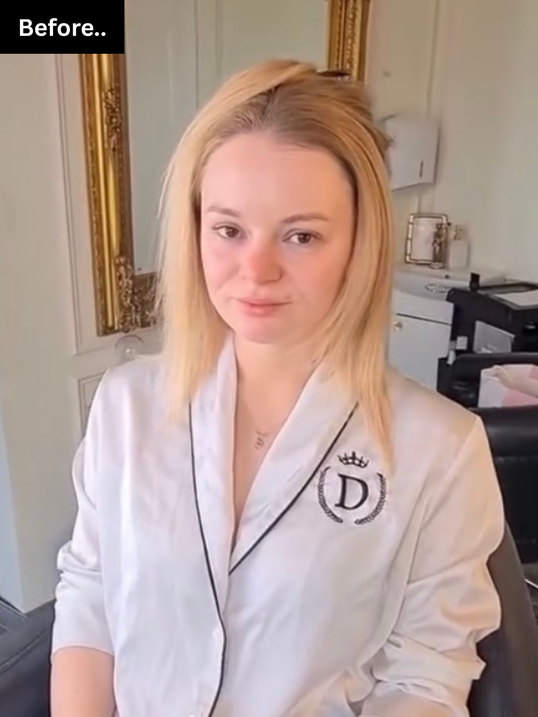 A young woman with blonde hair wearing a white satin robe with a black embroidered emblem and crown on the chest, standing in an indoor setting with a gold framed mirror and office equipment in the background.