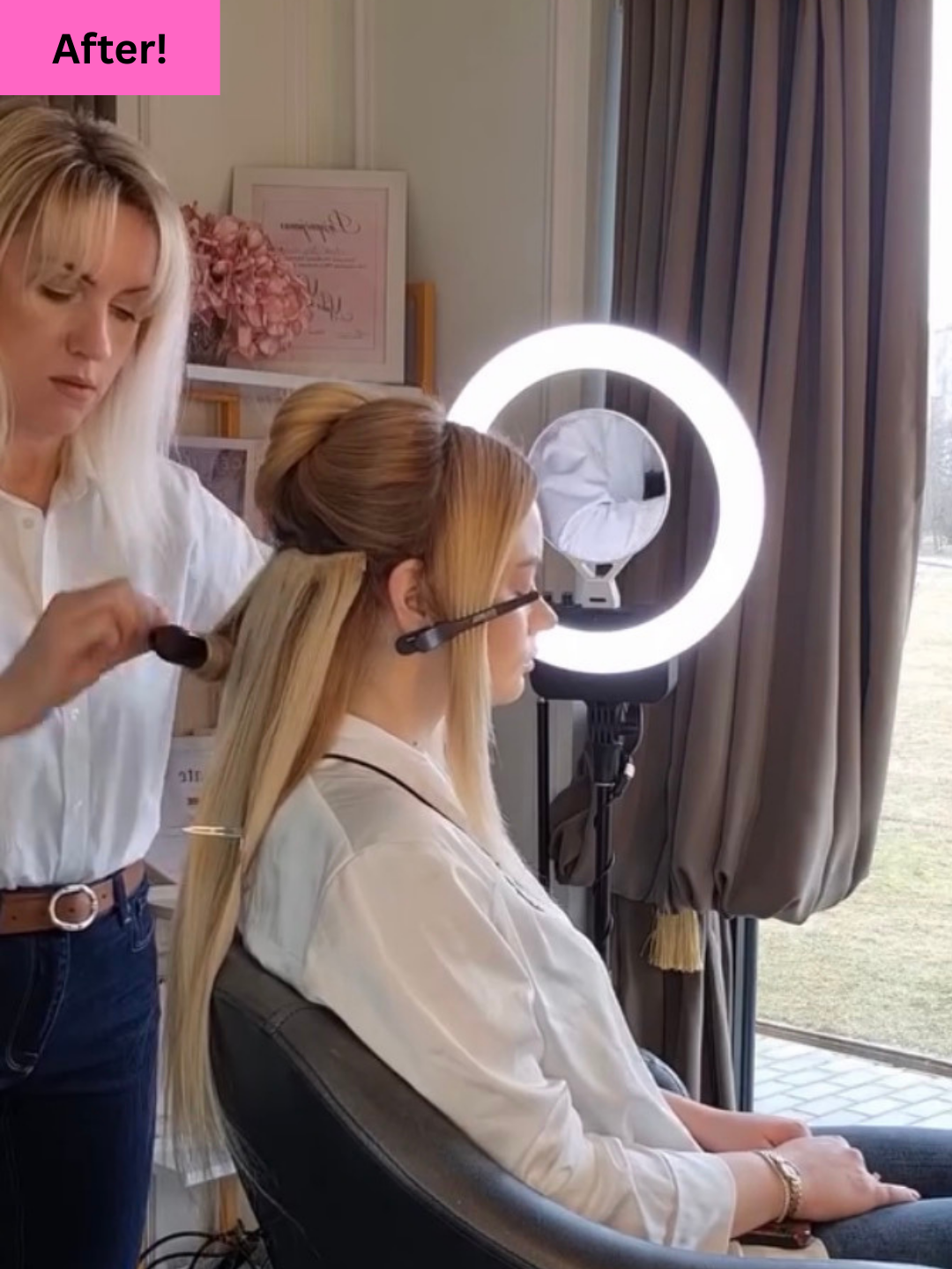 A woman sitting in a chair having her hair styled by a stylist, with a ring light, a mirror, and window with curtains in the background.