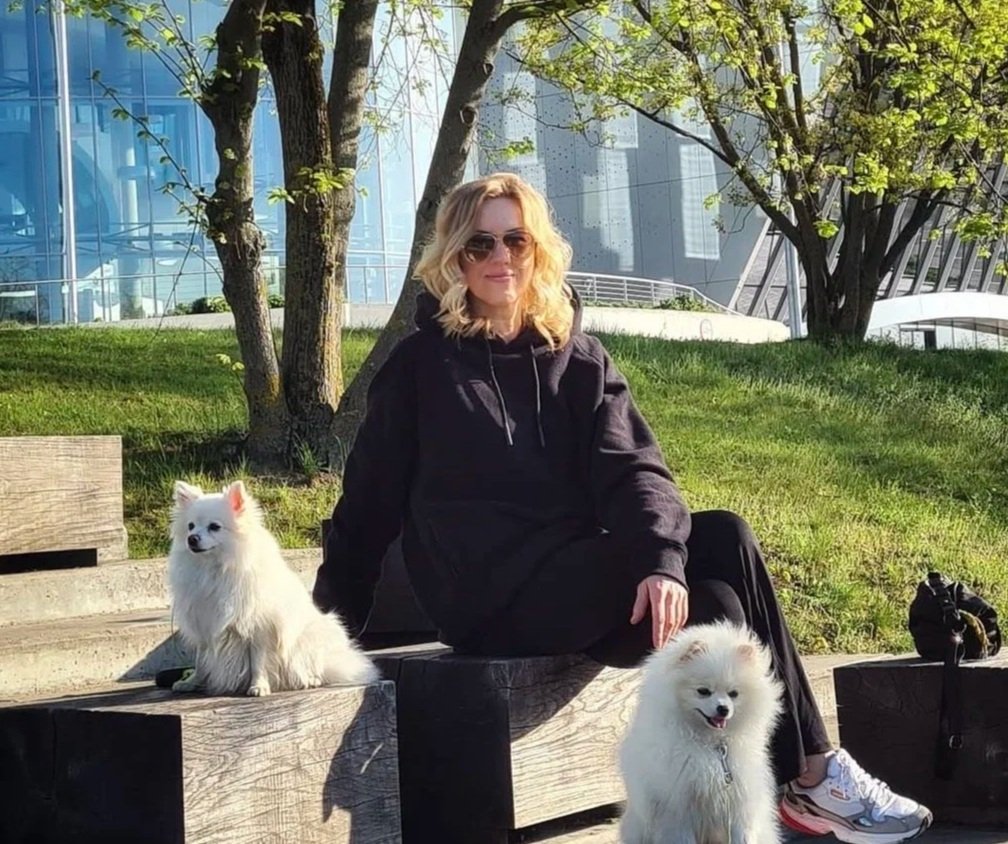 A woman sitting on a wooden bench in a park with two white fluffy dogs, one on each side. She is wearing sunglasses, a black hoodie, and black pants, with a modern building in the background.