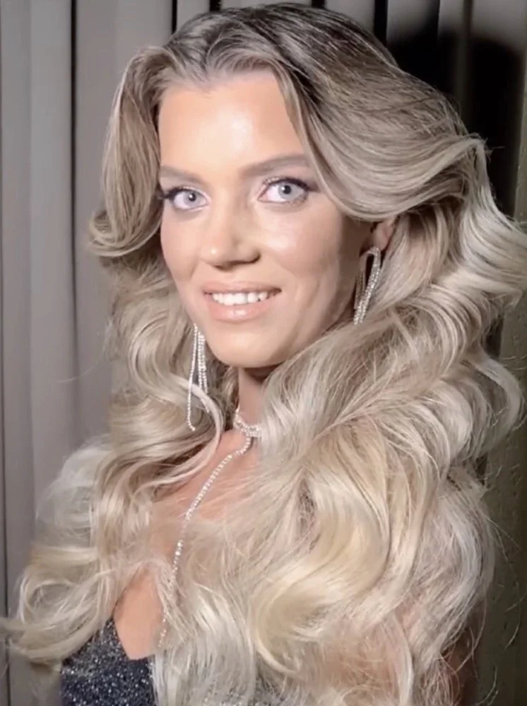 A woman with long, wavy platinum blonde hair, wearing sparkling earrings and a necklace, smiling for a portrait with a blurred background.