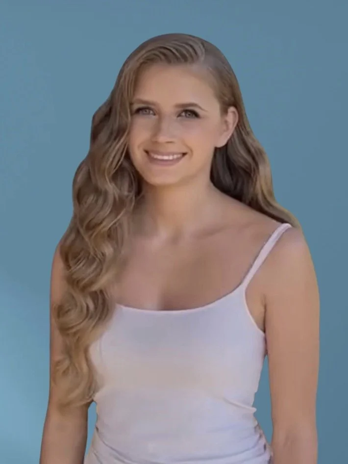 Portrait of a young woman with long, wavy light brown hair, smiling, wearing a white tank top against a solid blue background.