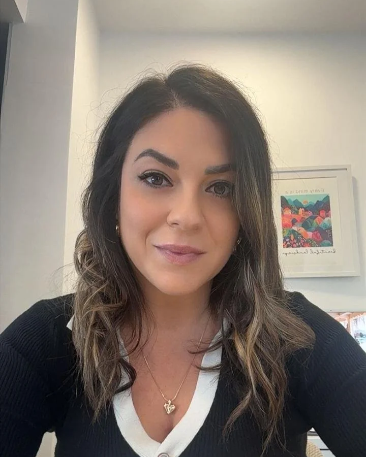 A woman with wavy brown hair and makeup, wearing a black top with a white collar, sitting indoors with framed artwork in the background.