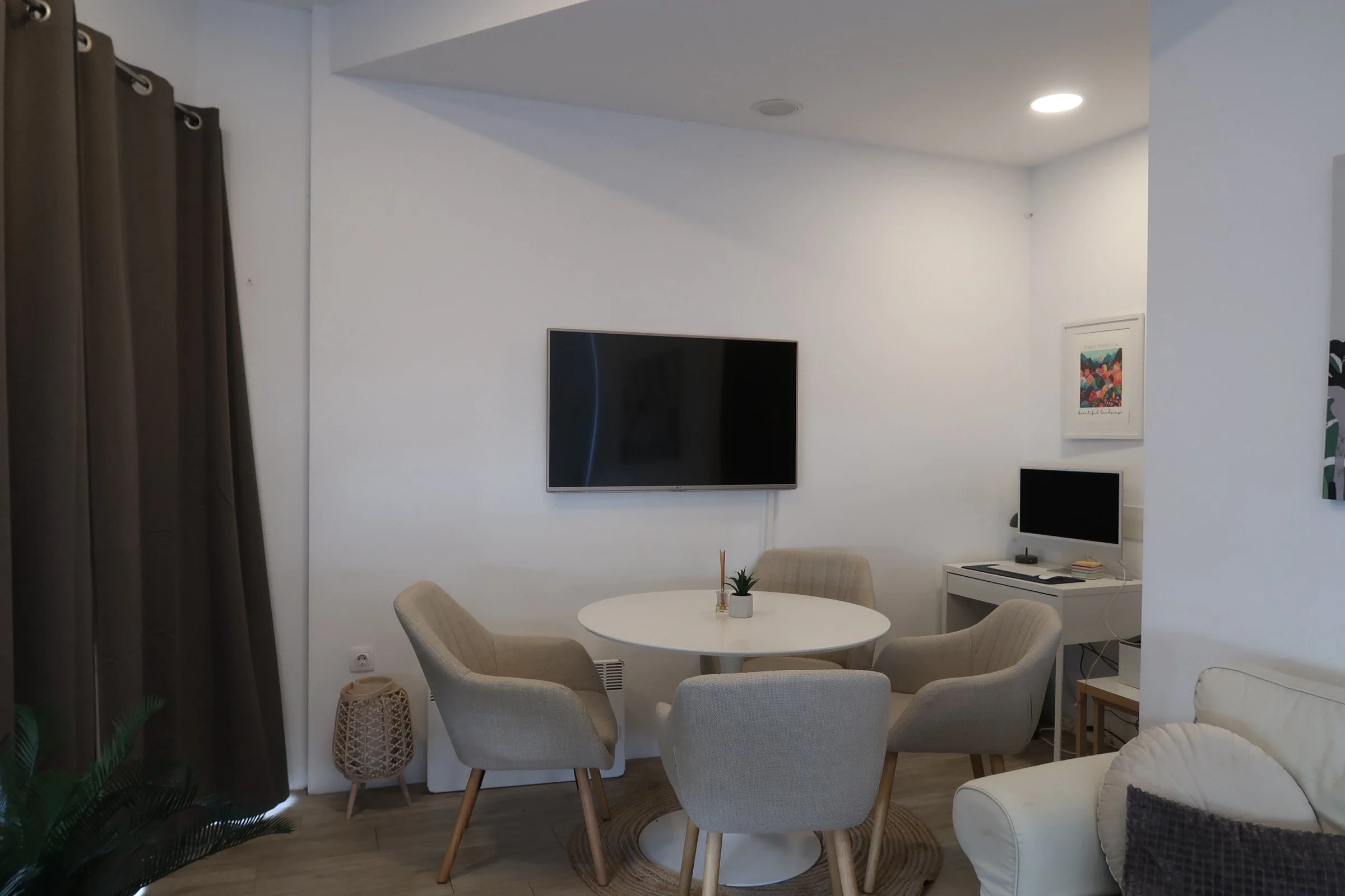 Living room with a white round table surrounded by six beige upholstered chairs, a wall-mounted flat-screen TV, a small desk with a computer monitor, and a beige sofa with a dark pillow, with dark curtains on the left and artwork on the right wall.