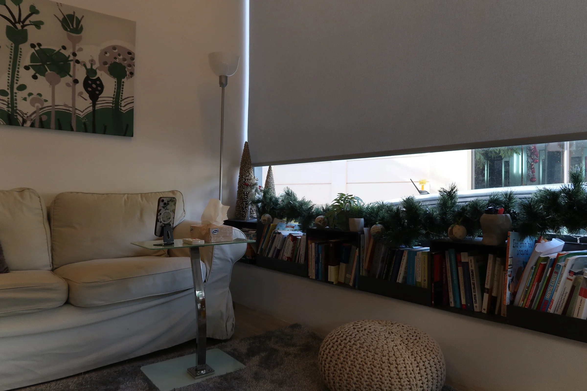 Living room corner with white sofa, small glass side table with phone, bookshelf with holiday decorations, window with partially closed blinds, and a knitted ottoman.