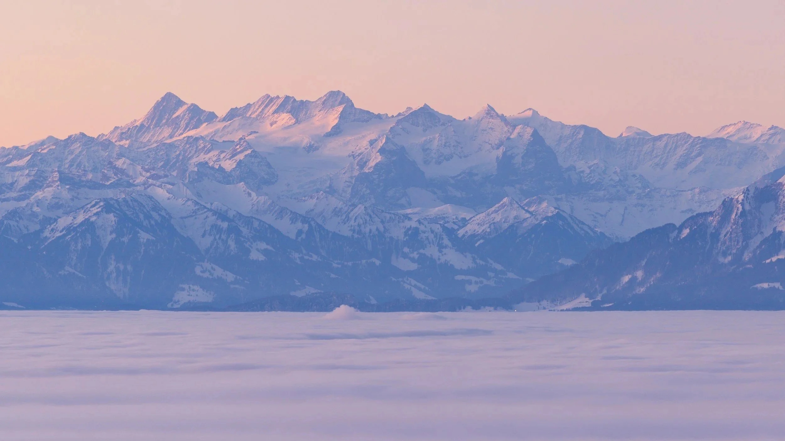Snow-covered mountain range with peaks in pinkish light, viewed over a frozen landscape.