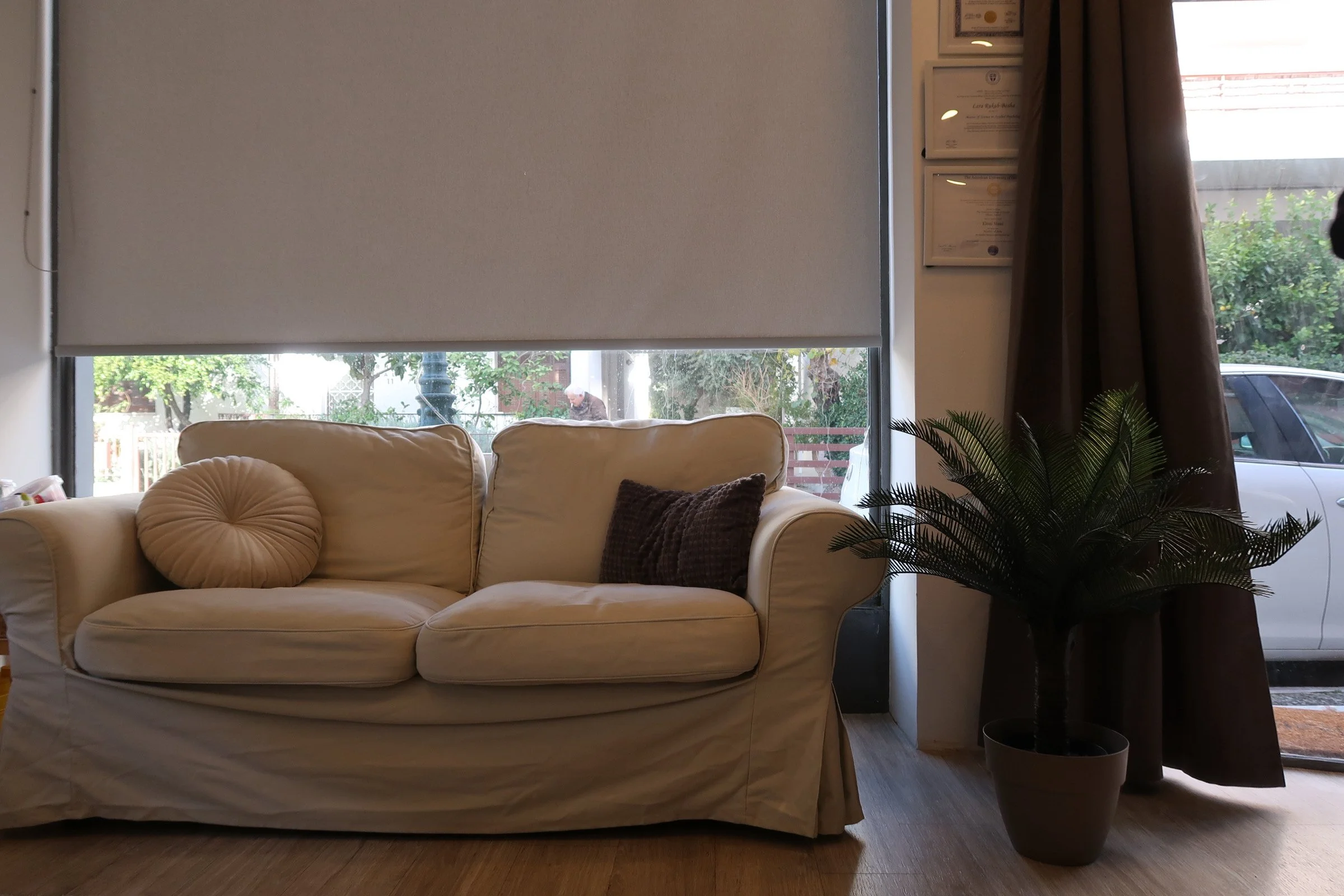 Interior of a room with a cream-colored sofa, brown and patterned pillows, a large window with a roller shade partially drawn, and a potted plant near the window, with curtains and framed certificates on the wall.