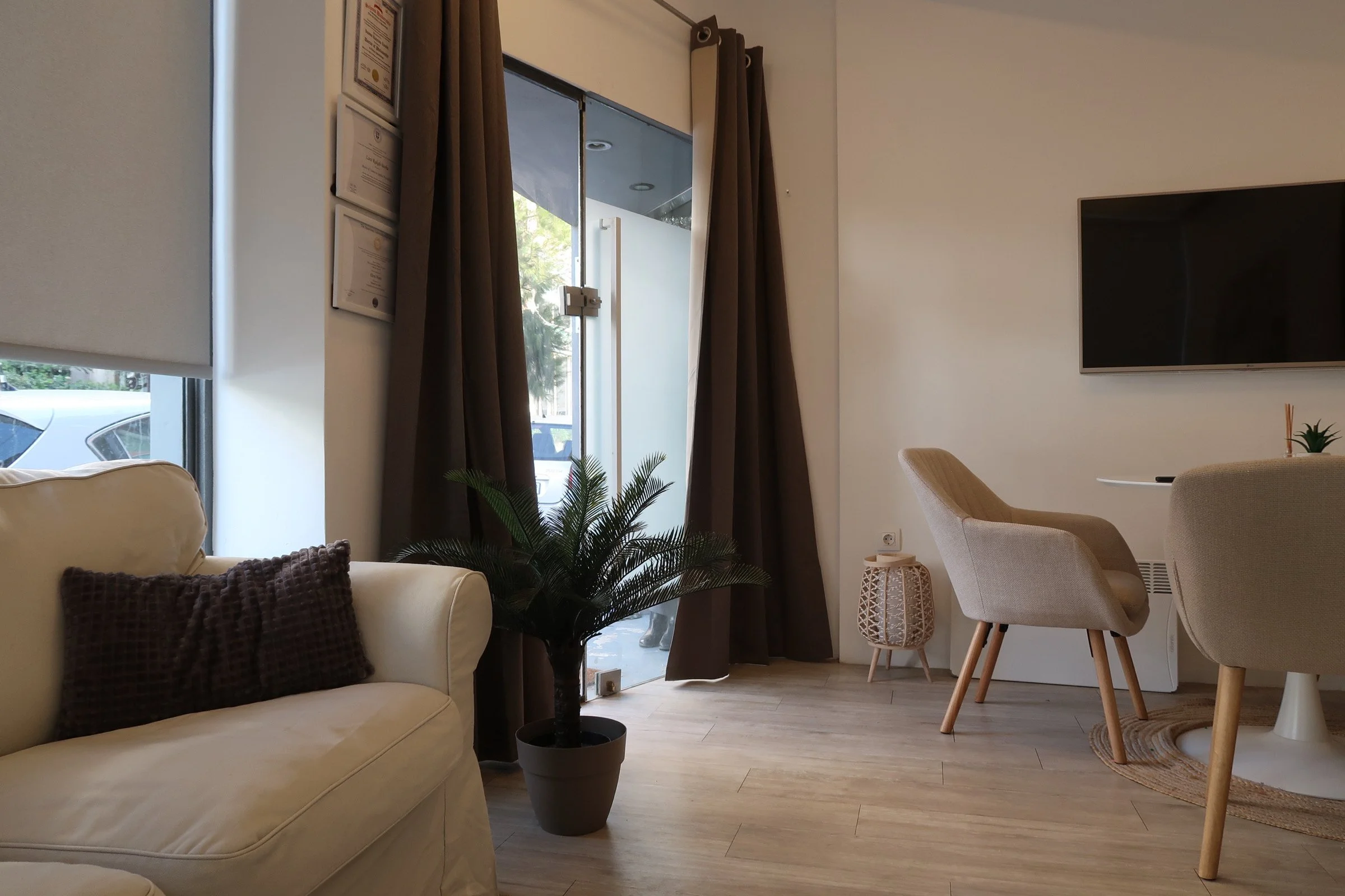 Cozy living room with cream-colored sofa, dark throw pillow, potted plant, large window with blinds and curtains, wall-mounted TV, beige chairs around a small table, and wooden flooring.