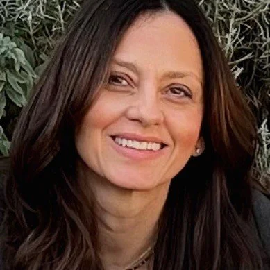 A woman with long brown hair smiling outdoors with greenery in the background.