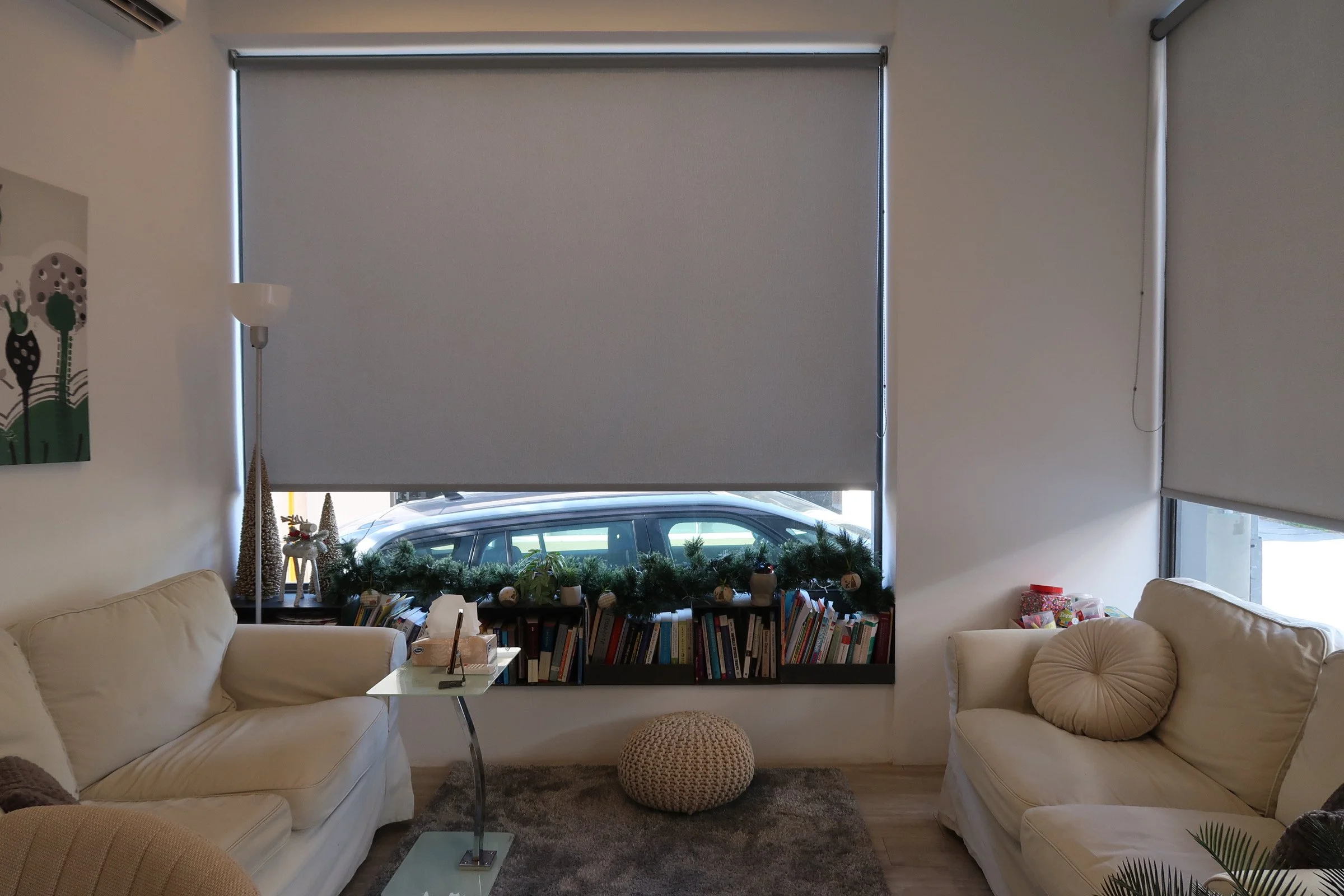 Living room with beige sofas, a glass coffee table, a knitted ottoman, a bookshelf with books and Christmas decorations, and windows with closed blinds.