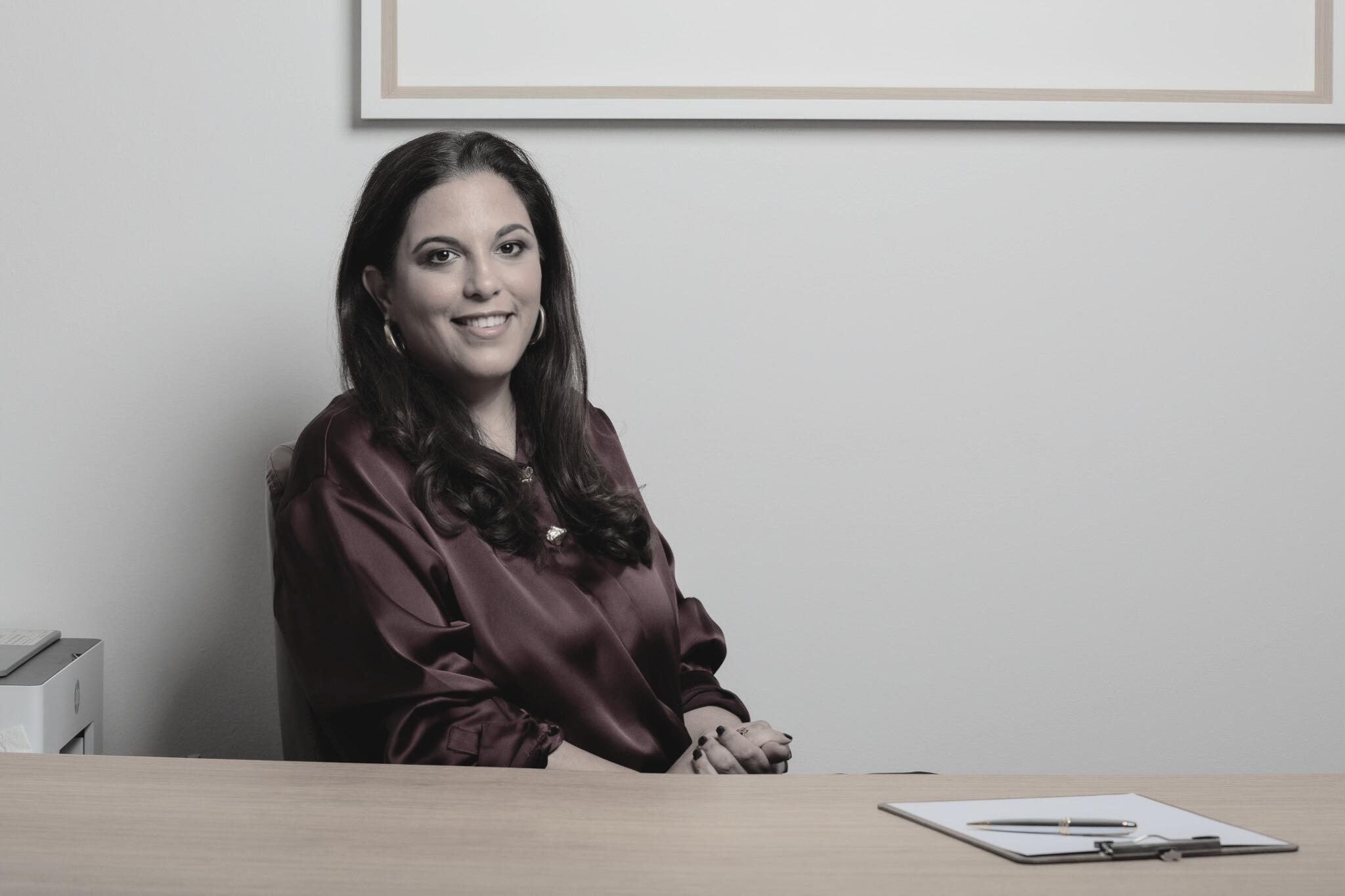 A woman with long brown hair, wearing a maroon satin blouse, smiling, sitting at a desk with a clipboard and pen, in an office setting with white walls and a framed picture above her.