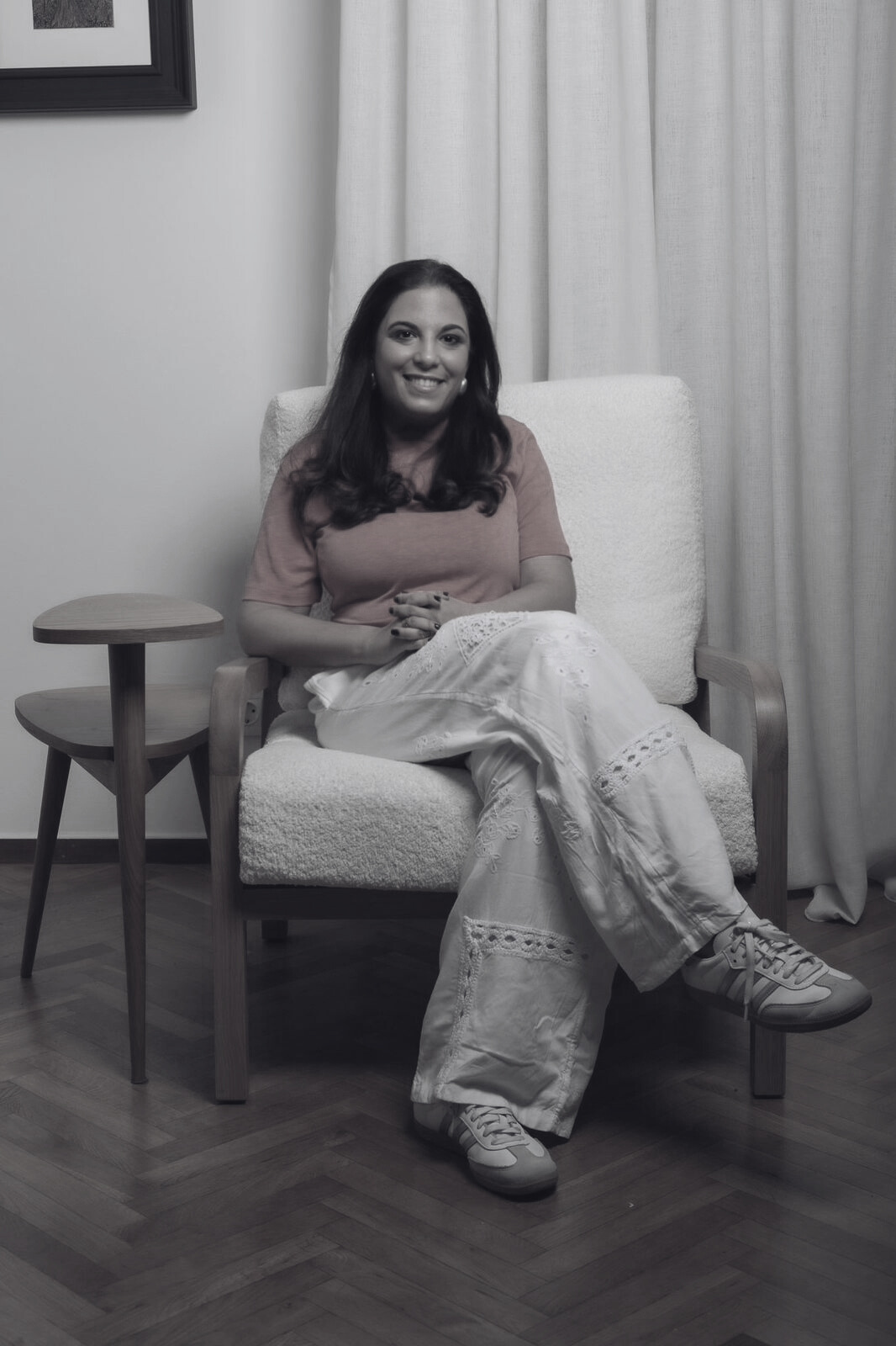 A woman sitting in a cream upholstered armchair in front of cream curtains, wearing a pink T-shirt, white embroidered pants, and sneakers, smiling at the camera.