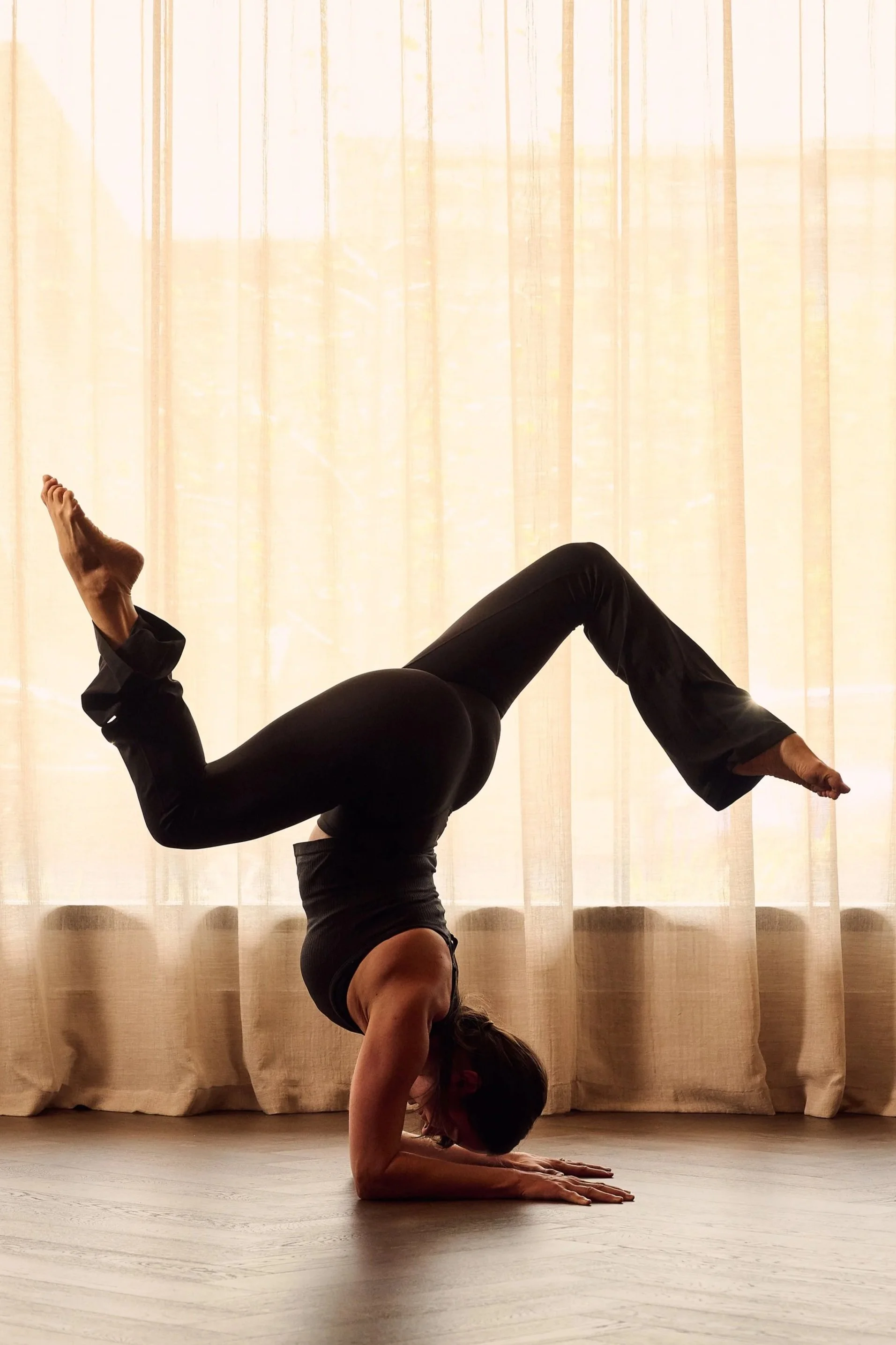 A woman doing a handstand with her legs bent at the knees in front of a large window with beige curtains, illuminated by natural sunlight.