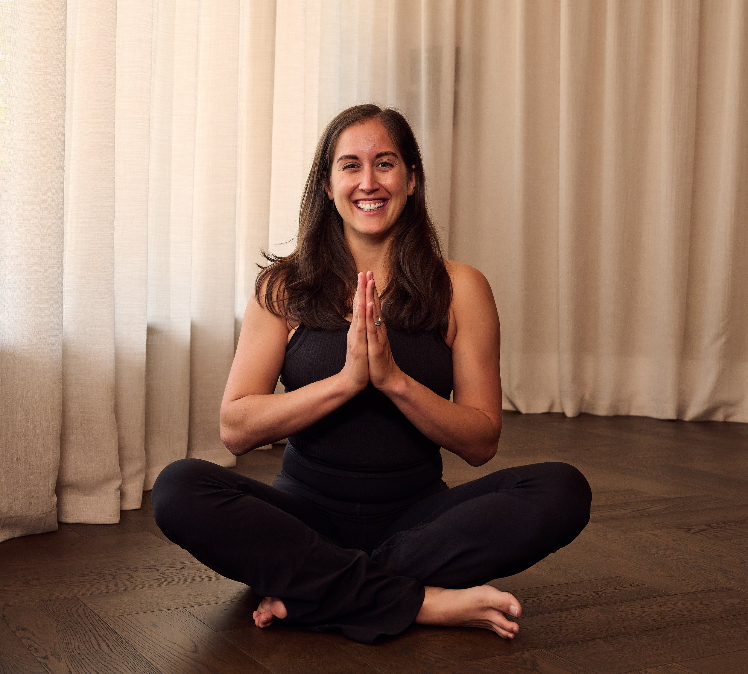 A woman sitting cross-legged on a wooden floor in front of beige curtains, smiling and holding her hands in a prayer position.