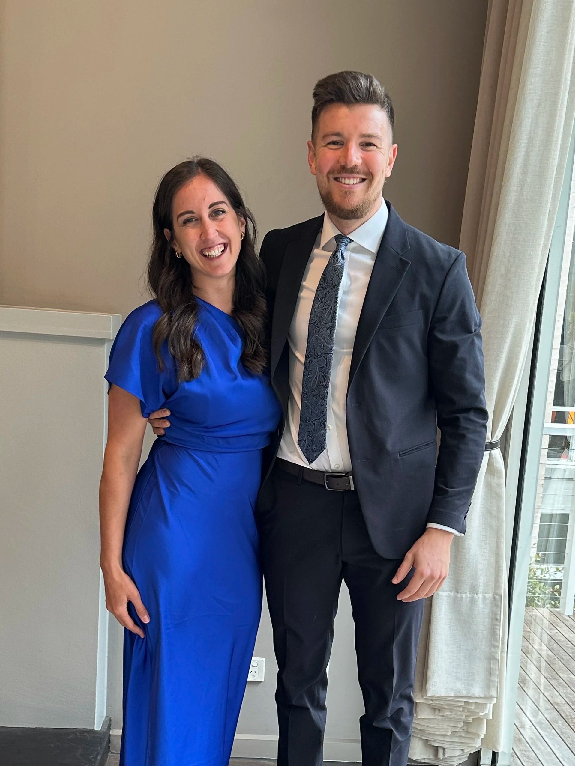 A man and woman dressed in formal attire smiling at the camera, standing close together indoors near a window with curtains.