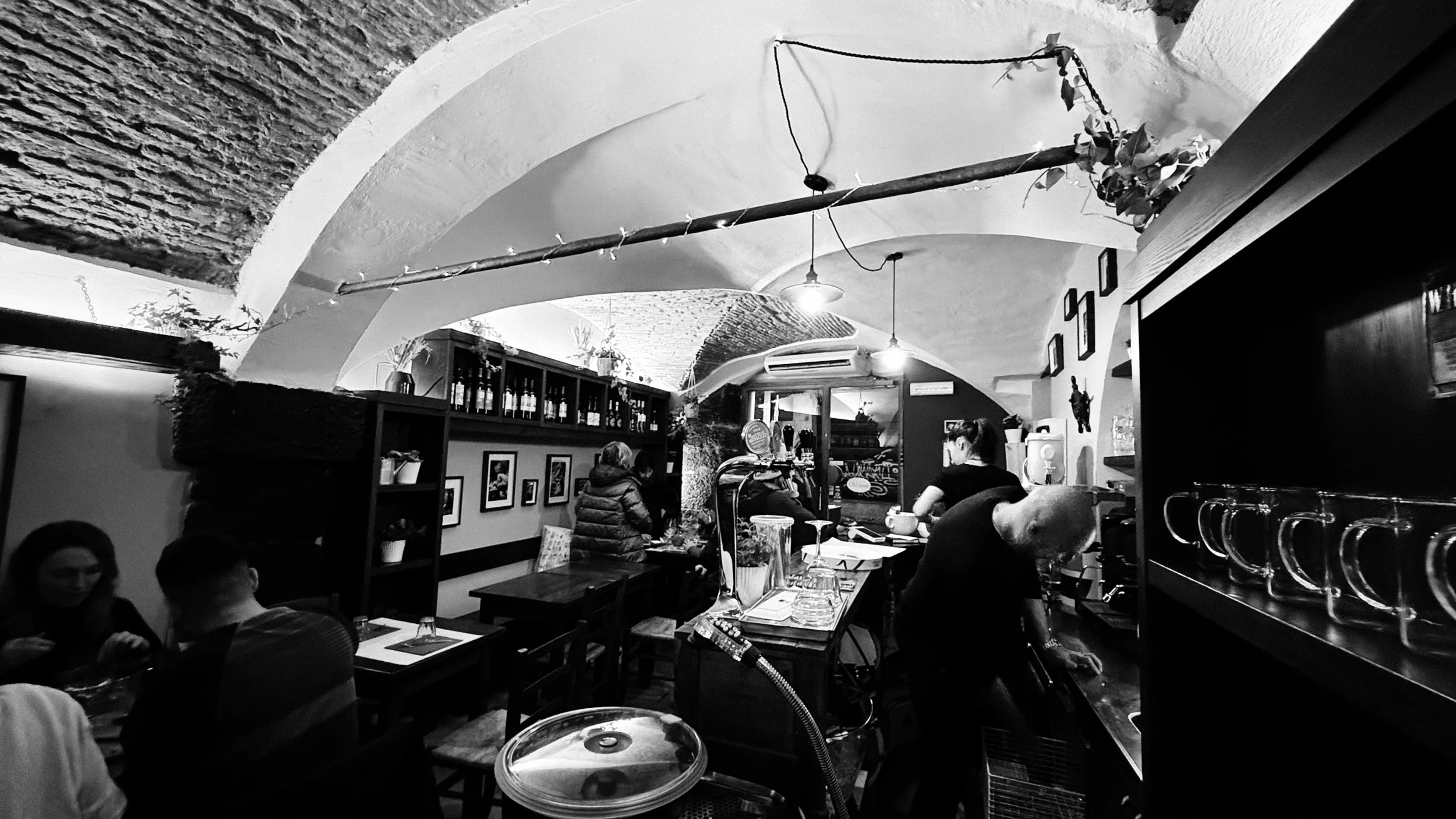 Interno di un ristorante o bar con archi in pietra, persone sedute ai tavoli, un barista che prepara una bevanda, scaffali con bottiglie e ornamenti, pareti decorate con quadri e oggetti. L'ambiente è arredato in stile accogliente e rustico.