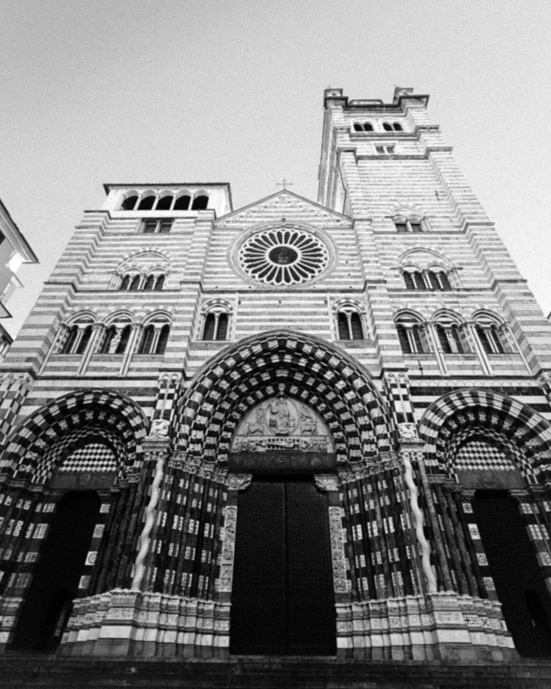 Vista frontale di una chiesa medievale con facciata in pietra con motivi geometrici in bianco e nero e un rosone centrale, due torri ai lati e un grande portale d'ingresso.