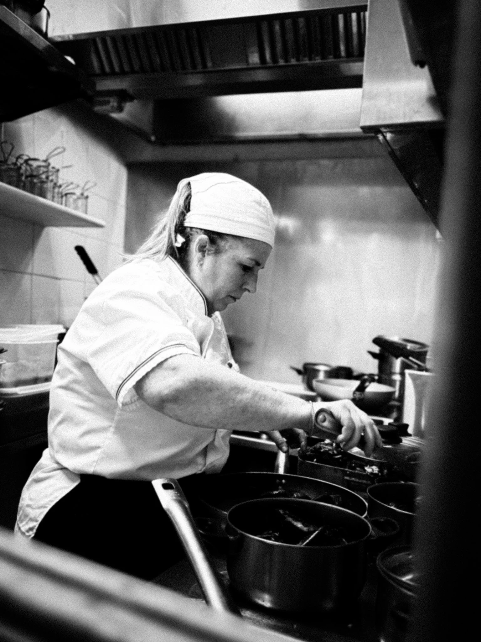 Cuoca che cucina in una cucina professionale, indossa un cappello e una divisa, mentre lavora con pentole e utensili.