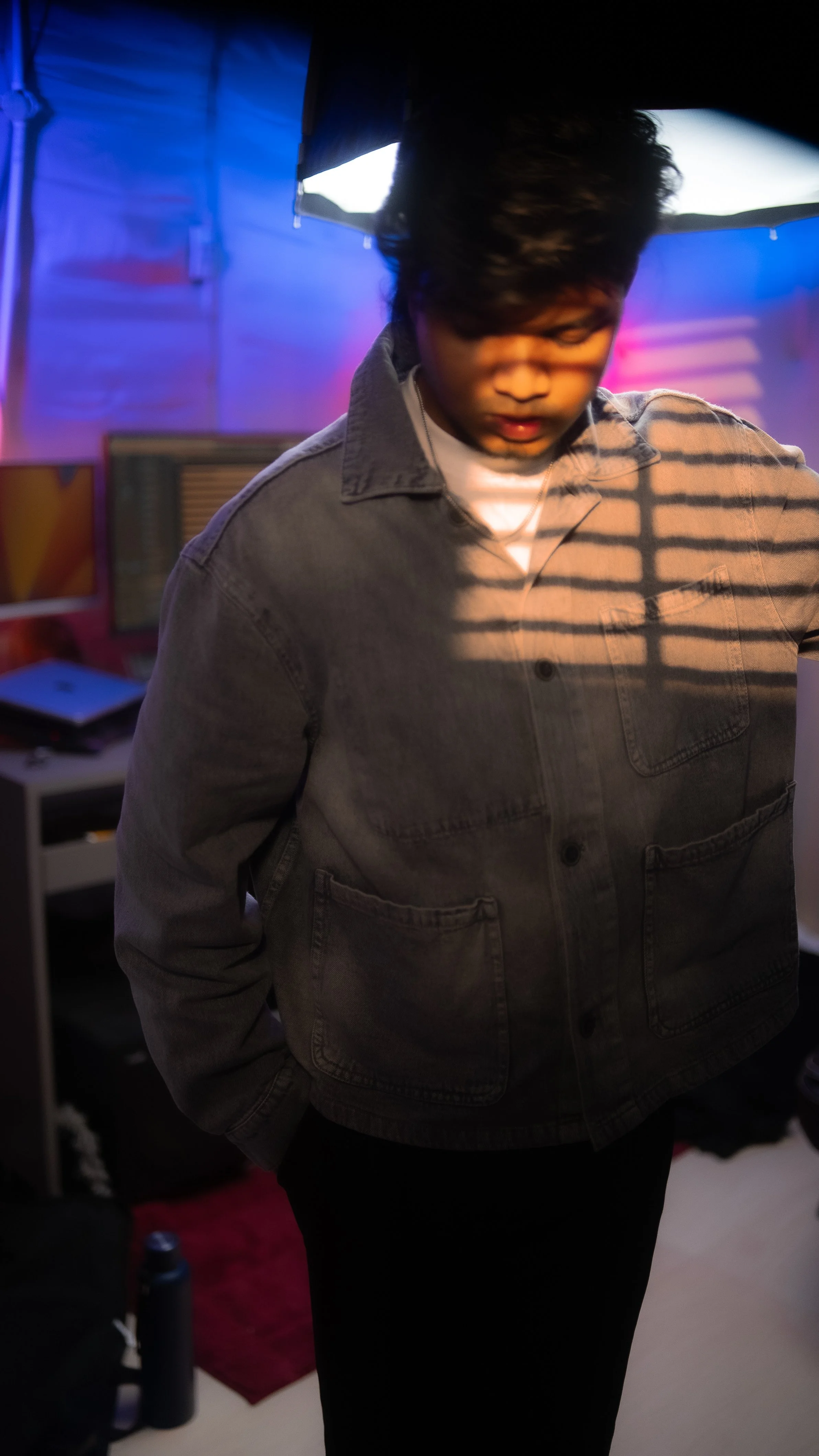 A young man with dark hair wearing a denim jacket, looking down, in a room with colorful ambient lighting and a computer behind him.