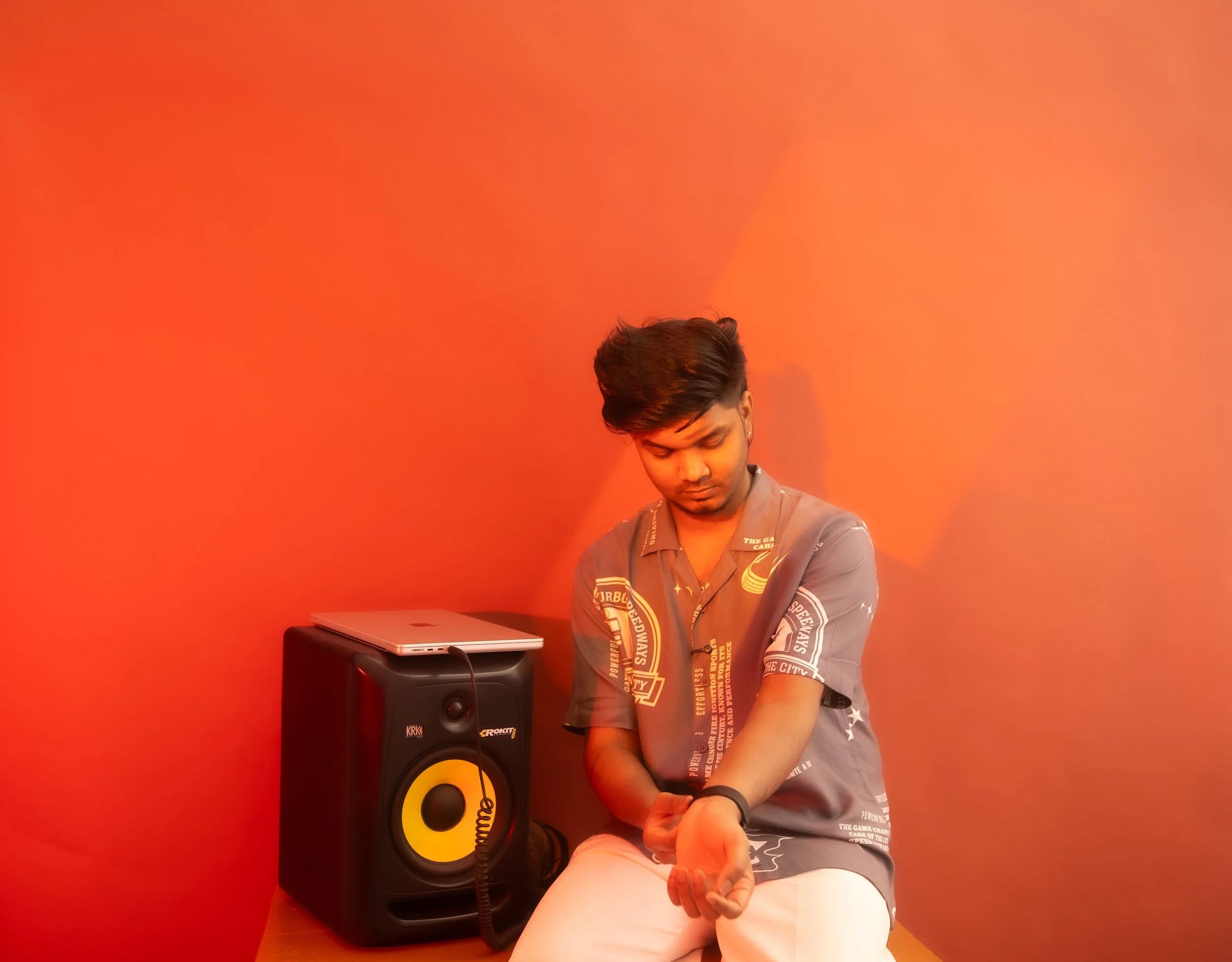 A young man sitting against a coral-colored wall, with a speaker and a closed laptop on top of the speaker, looking down and adjusting his sleeve.