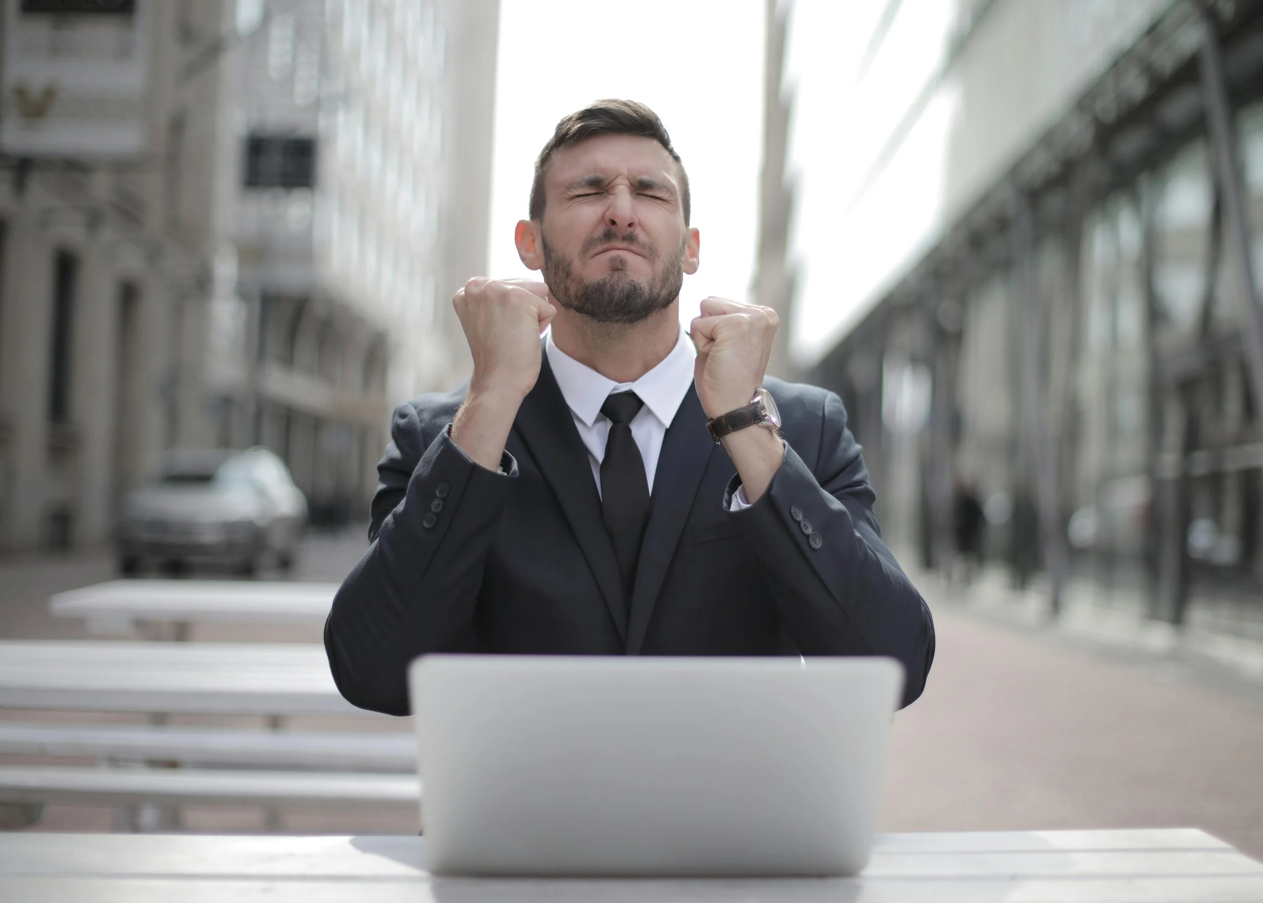 Businessman in a suit showing a victorious gesture with clenched fists while sitting at a table with a laptop outdoors.