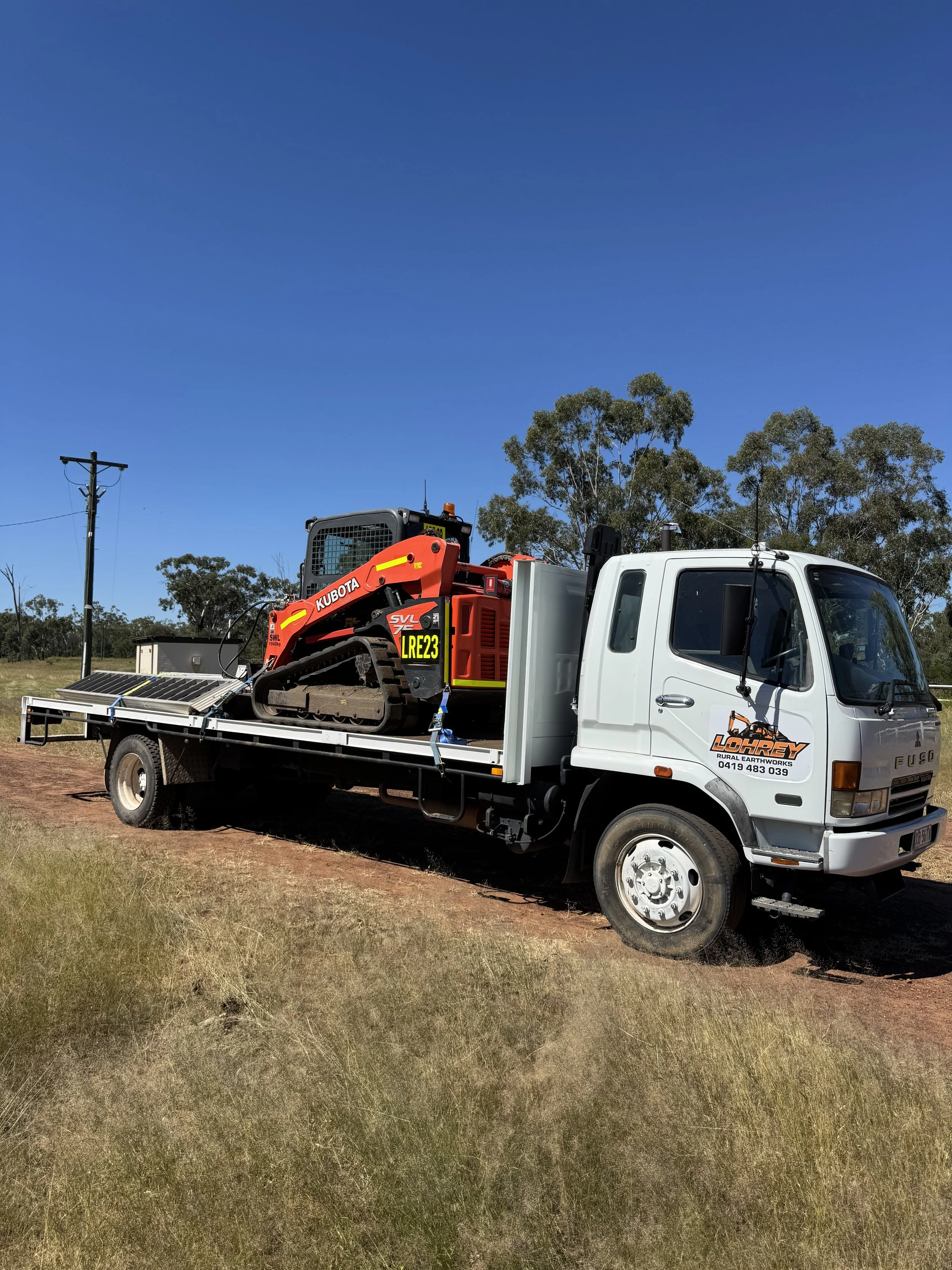A white flatbed truck parked on grass with a small red and black tracked earthmoving machine on the back, under a clear blue sky.