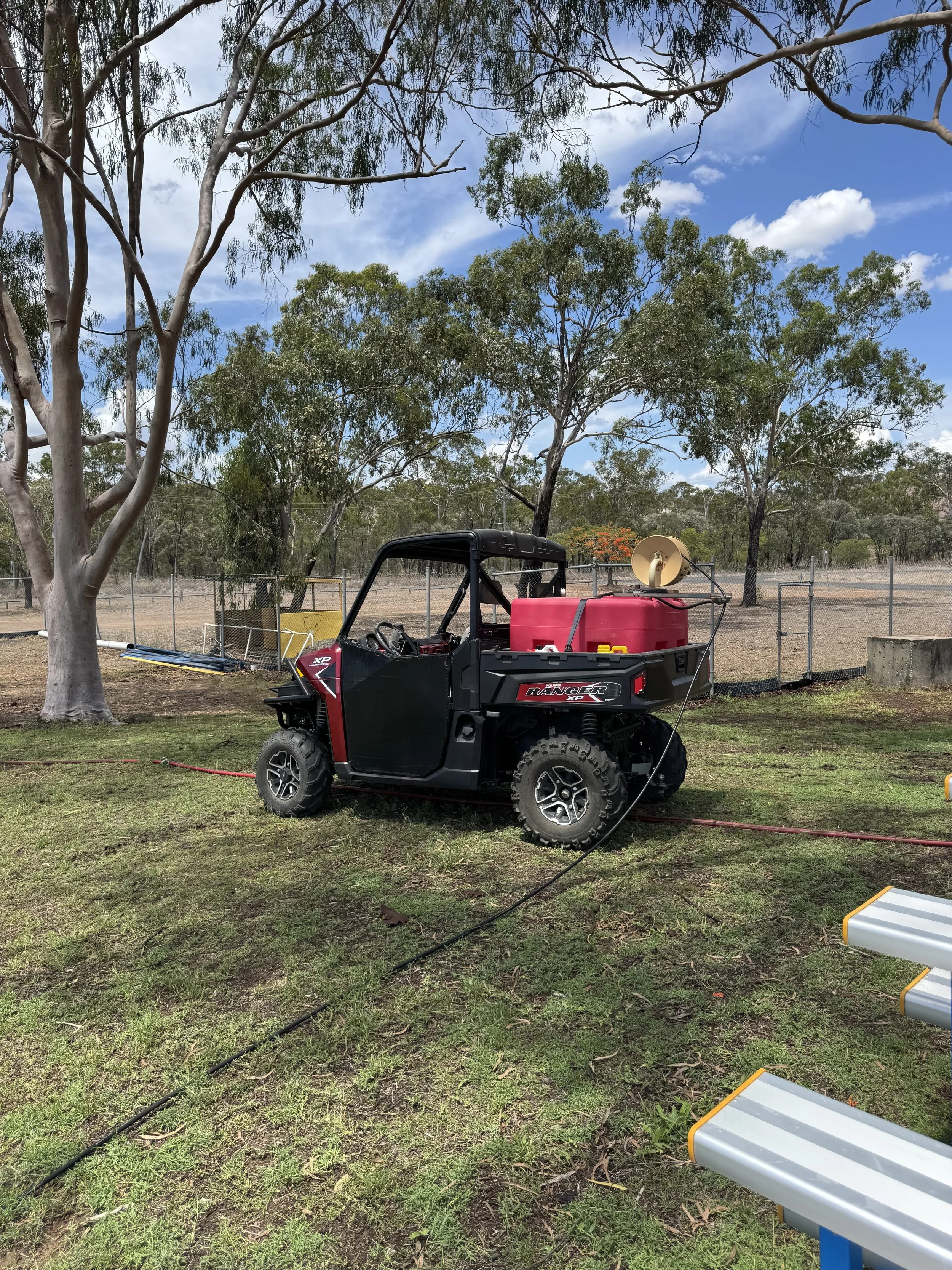 A black and red Utility Terrain Vehicle (UTV) parked on grass, with a red spray tank on the back, in an outdoor area with trees and a blue sky with clouds.