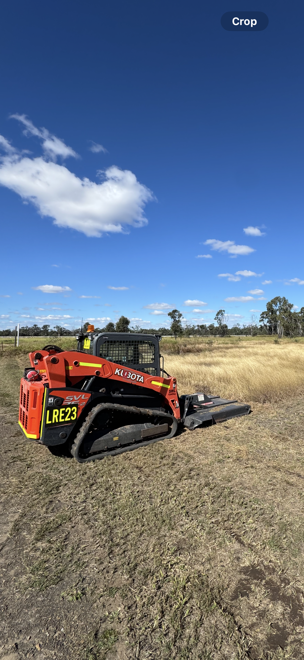 A Kubota compact tracked skid steer loader with yellow and black notification stickers in a grassy field under a blue sky with scattered clouds.