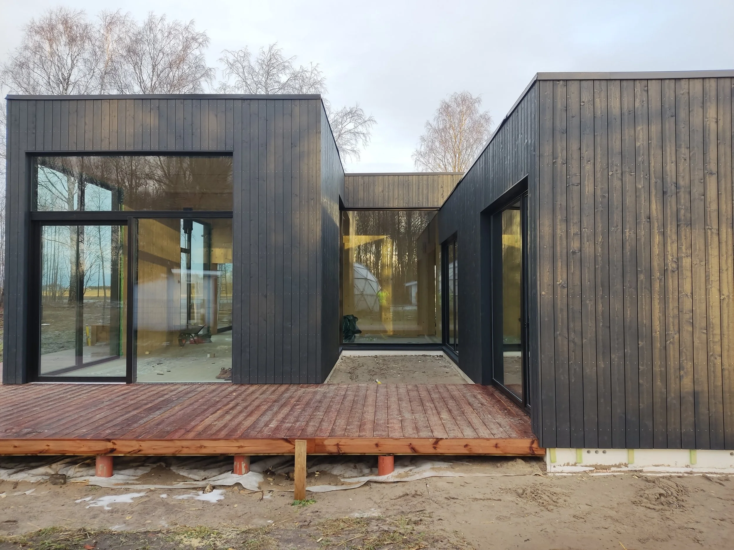 Värskelt valminud modernne maja musta puitfassaadi, suurte klaasakende ja klaasuksega. Maja ees on väike puitterrass, ümbrus on avatud ja valgusküllane – ideaalne koht, kus terrassil hommikukohvi juua.