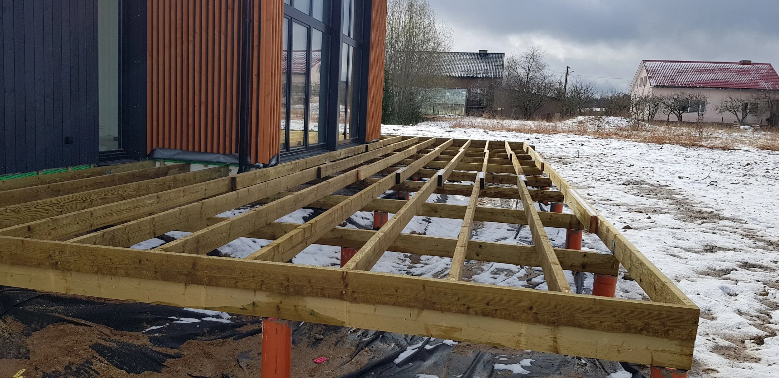 Puitterrassi ehitus kaasaegse, suurte klaasakendega maja ette lumisel talvepäeval. Terrassi karkass on pooleli, ümberringi on lumi – töö käib, et luua mugav väliruum ka põhjamaistes tingimustes.