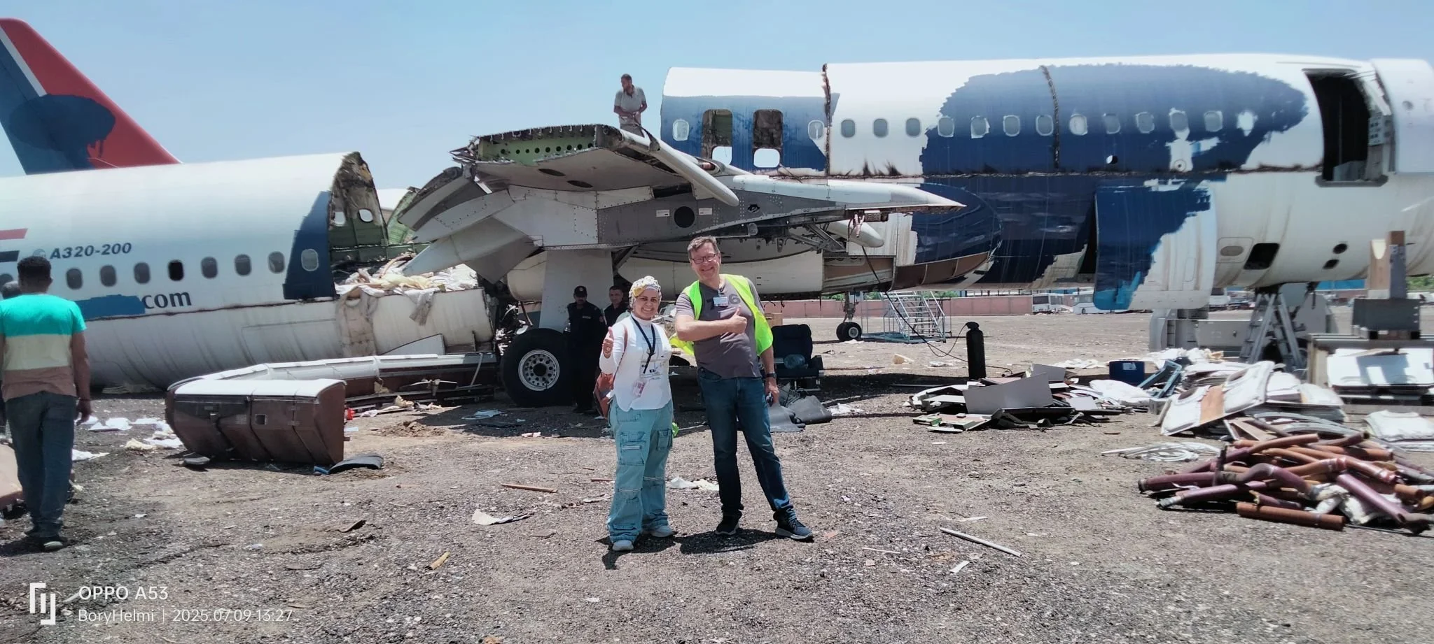 Two smiling people standing in front of a crashed airplane, giving a thumbs-up, with debris around and damaged aircraft parts nearby.