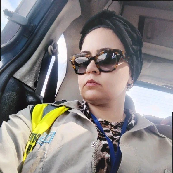 Woman with short dark hair wearing large sunglasses, a headscarf, and a beige uniform with a bright yellow and black strap, sitting inside a vehicle.