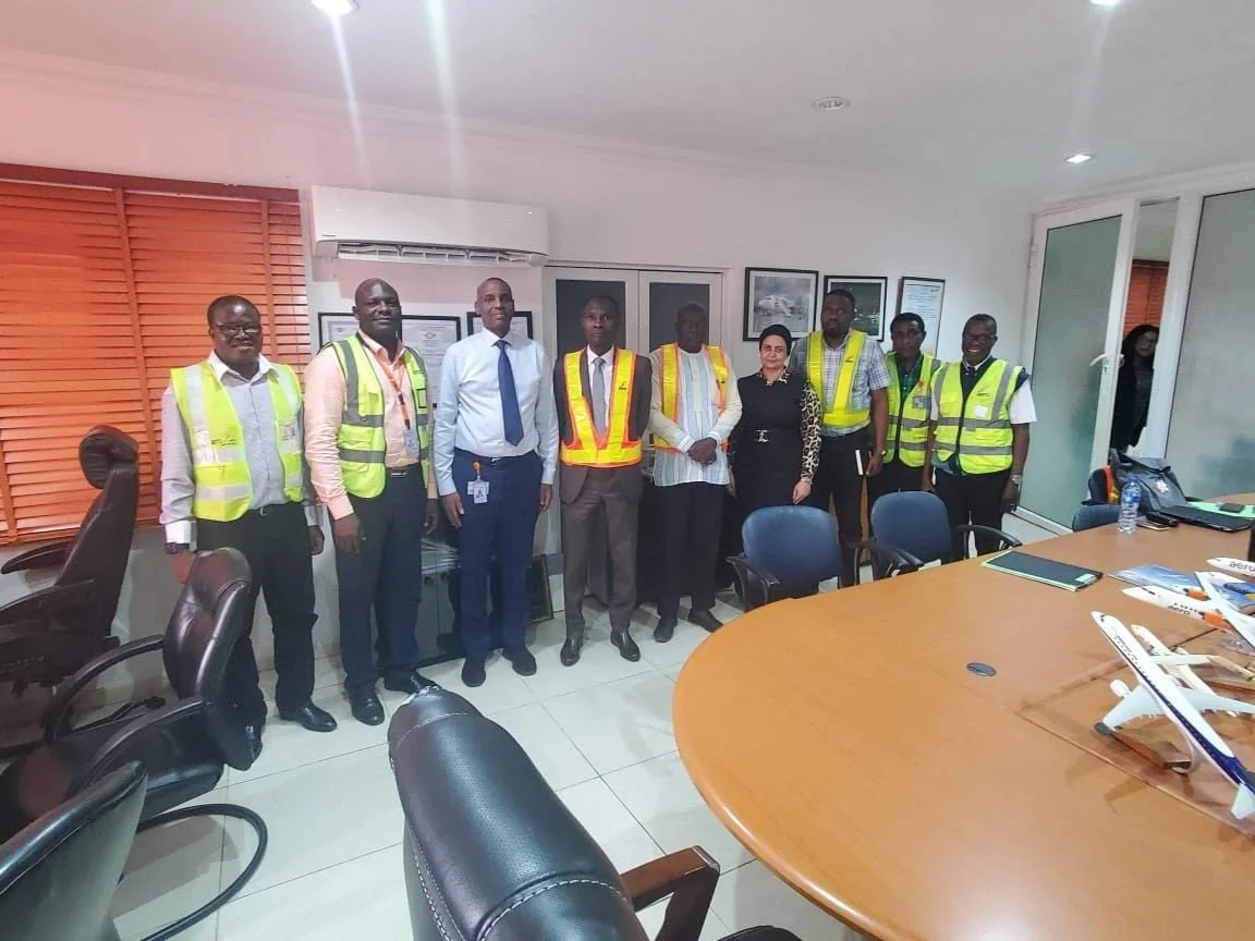 Group of eight people in safety vests standing in a conference room with a large oval table, chairs, and model airplanes on the table.