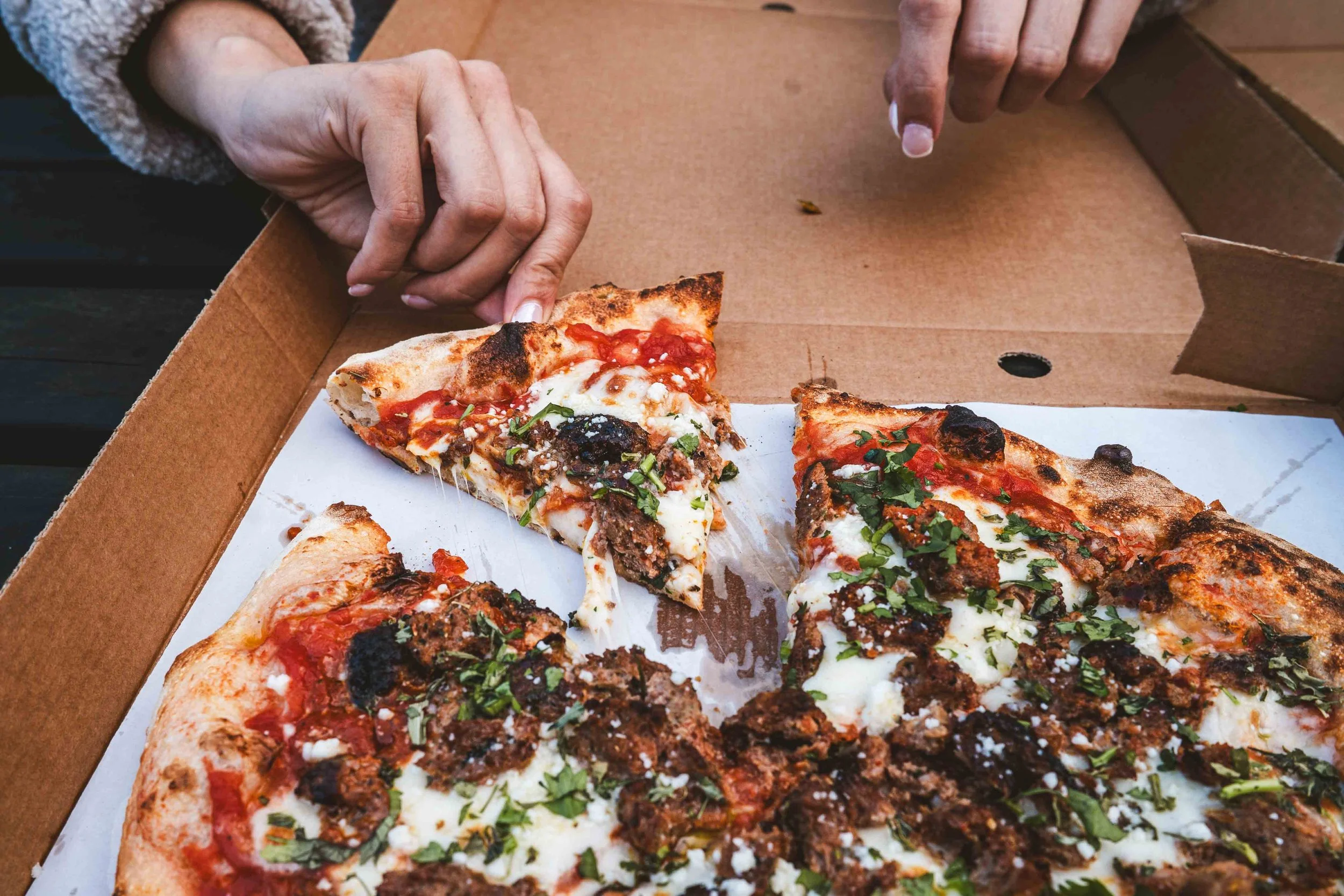 Hand taking a slice of Lamb Keema Sizzla fusion pizza from a Curry Town takeaway box, showcasing the stonebaked crust and spiced halal lamb toppings in Crawley