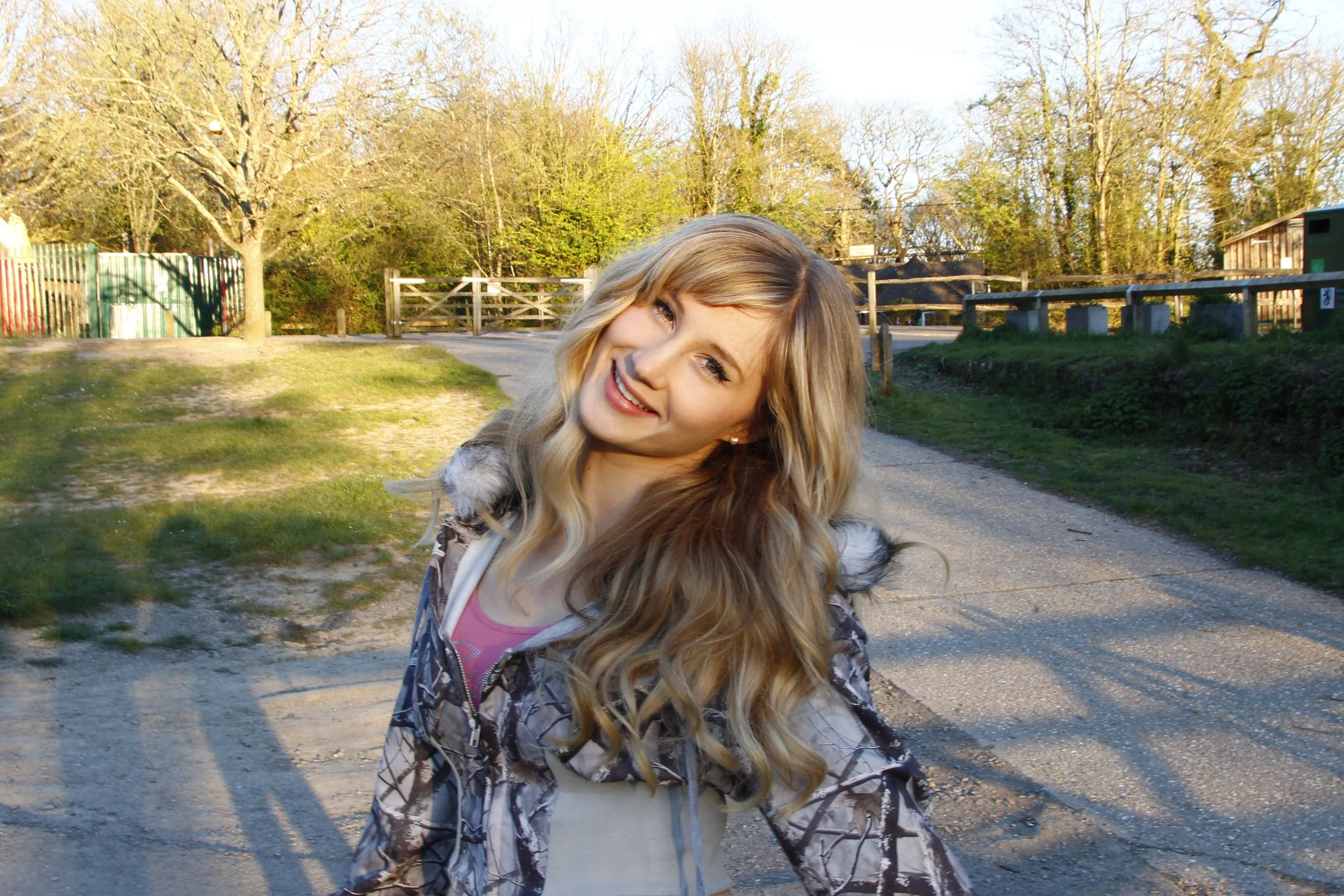 A young woman with long blonde hair smiling outdoors in a park or nature area during daytime.