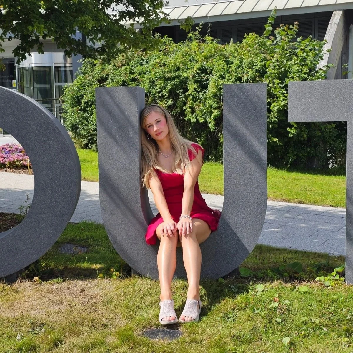 A young woman with long blonde hair in a red dress and heels sitting on a large gray letter sculpture outdoors, with green bushes and a building in the background.