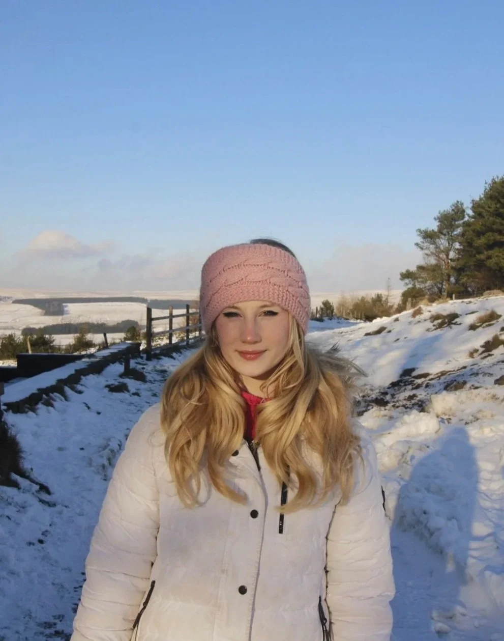 A young woman standing outdoors in a snowy landscape, wearing a pink knit hat and a white winter coat, with a clear blue sky and some trees in the background.