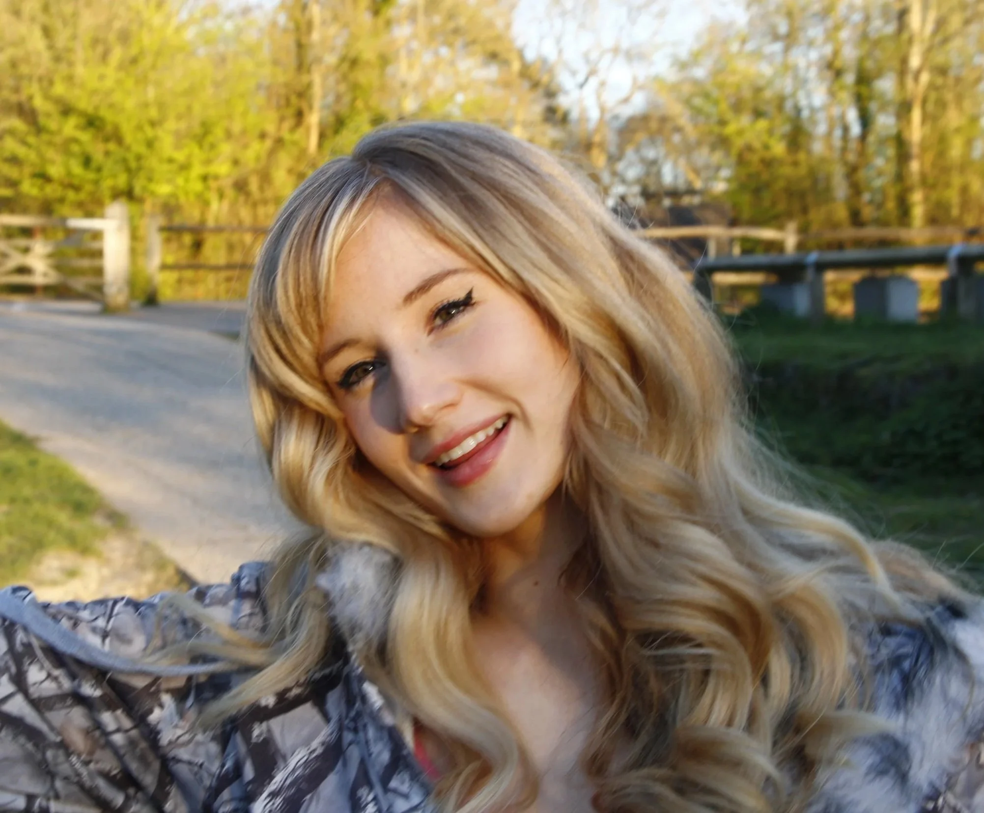 A woman with long blonde hair smiling outdoors on a sunny day, standing on a dirt path with trees and a wooden fence in the background.