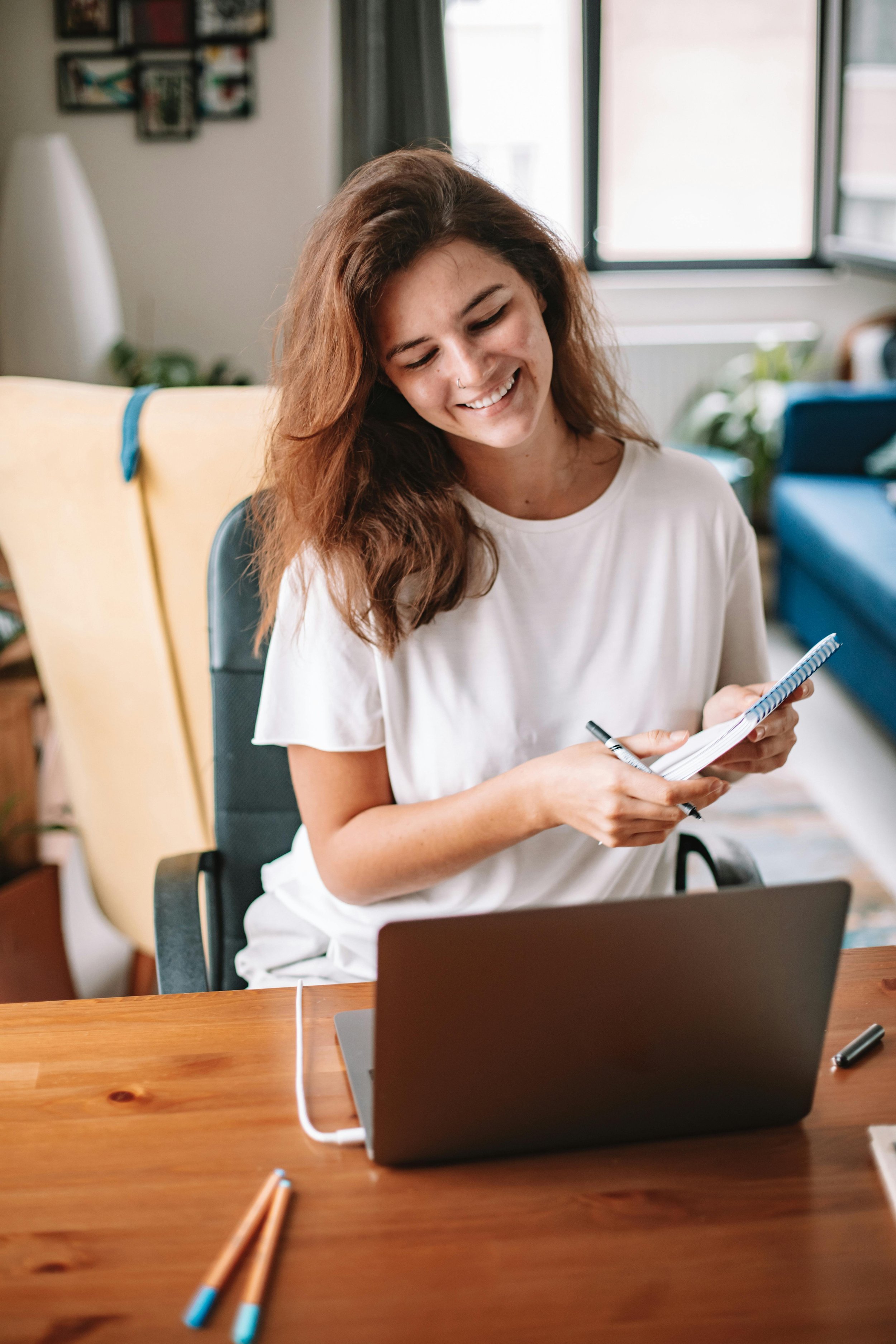 A woman with shoulder-length brown hair smiling, sitting at a wooden desk, holding a notepad and pen, with a laptop open in front of her in a cozy, well-lit room.