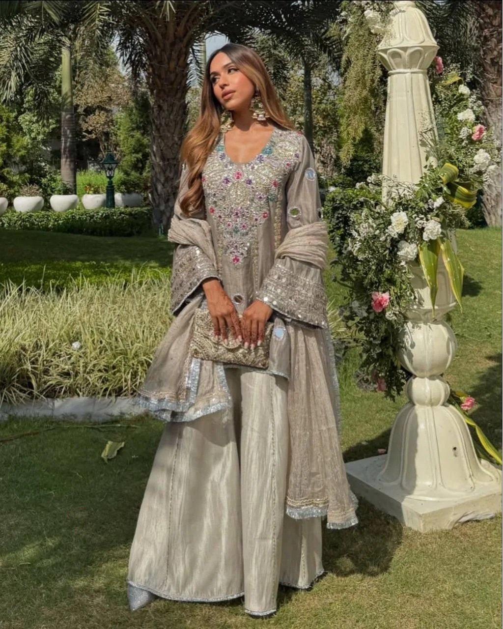 A woman in an elegant beige traditional outfit with intricate embroidery and embellishments, standing outdoors in a garden next to a large floral arrangement in a white pedestal.