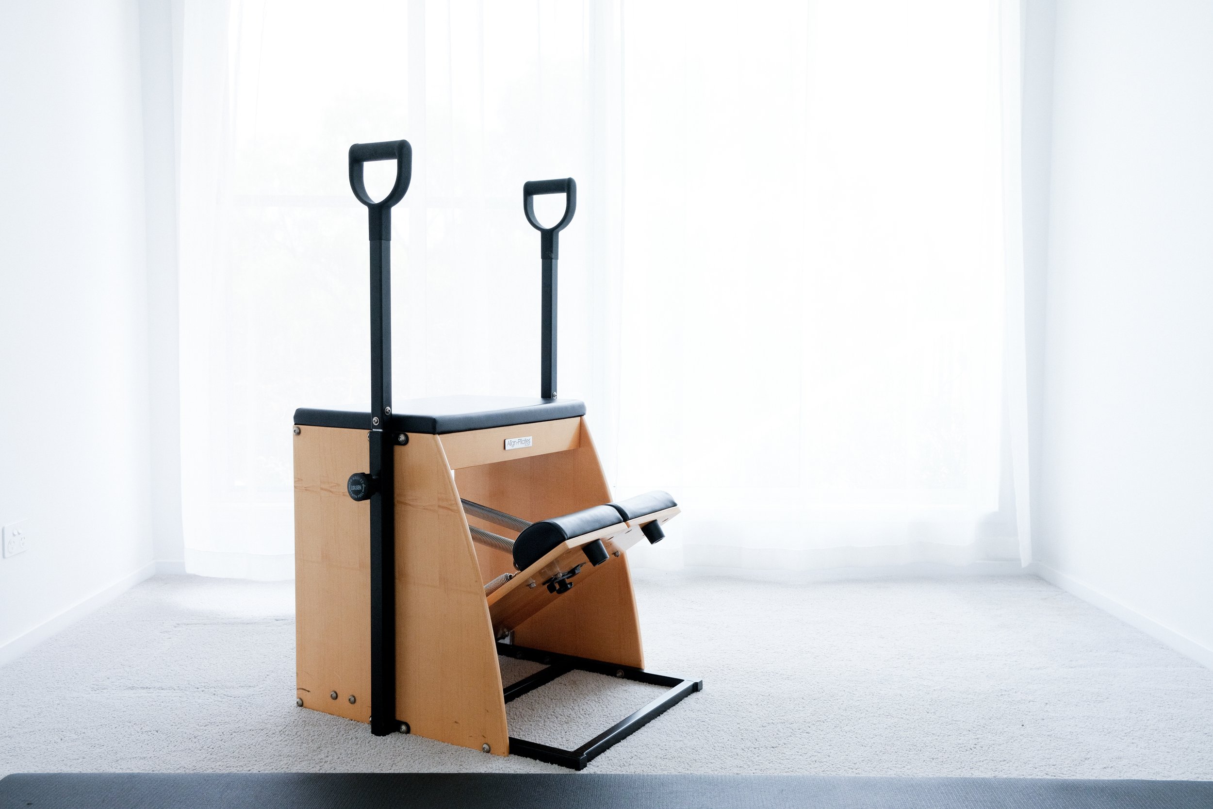 A Pilates chair, a piece of exercise equipment made of wood and black metal, placed on a light-colored carpet in a room with sheer white curtains allowing natural light.