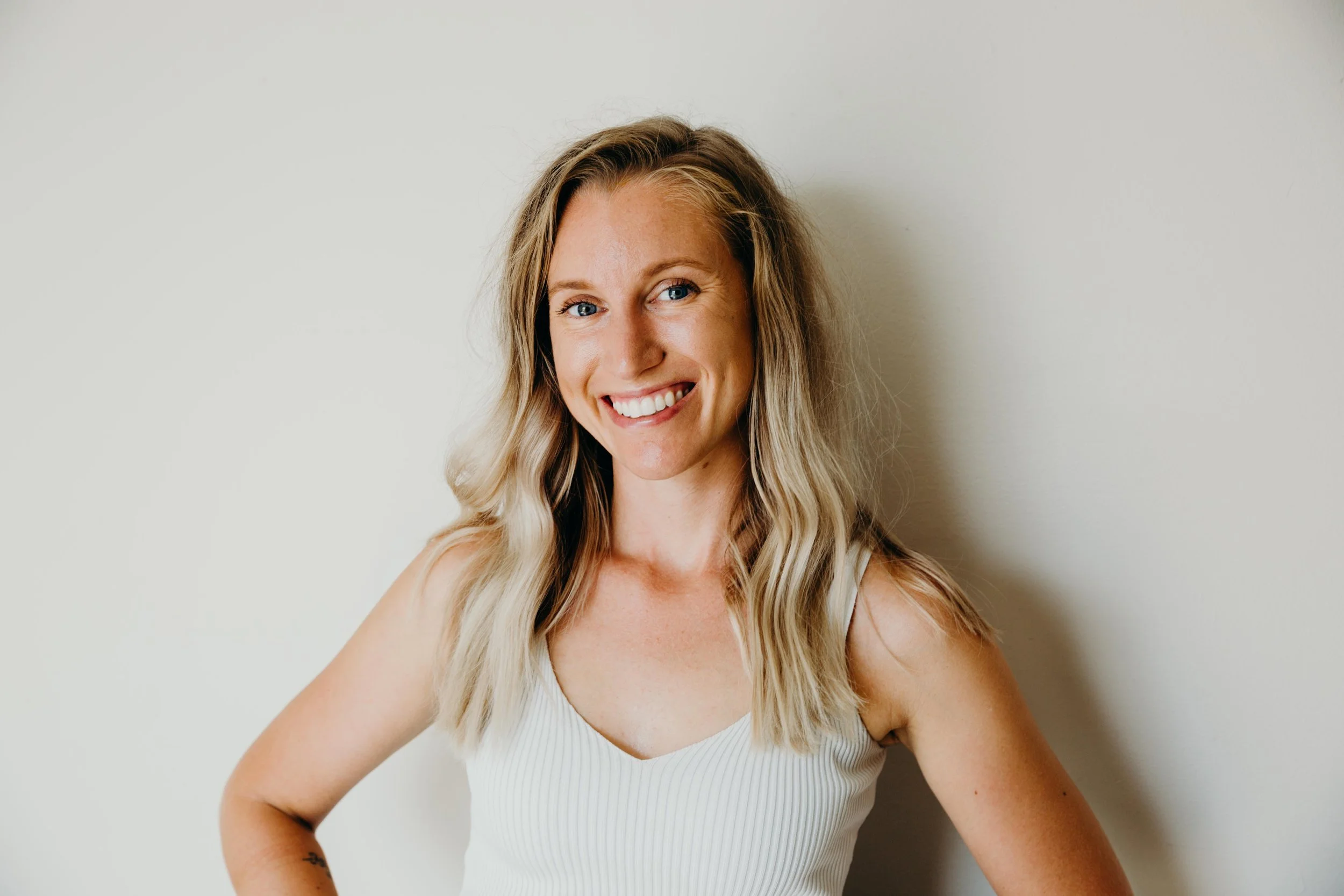 A woman with blonde hair, blue eyes, and a broad smile standing against a plain white wall.