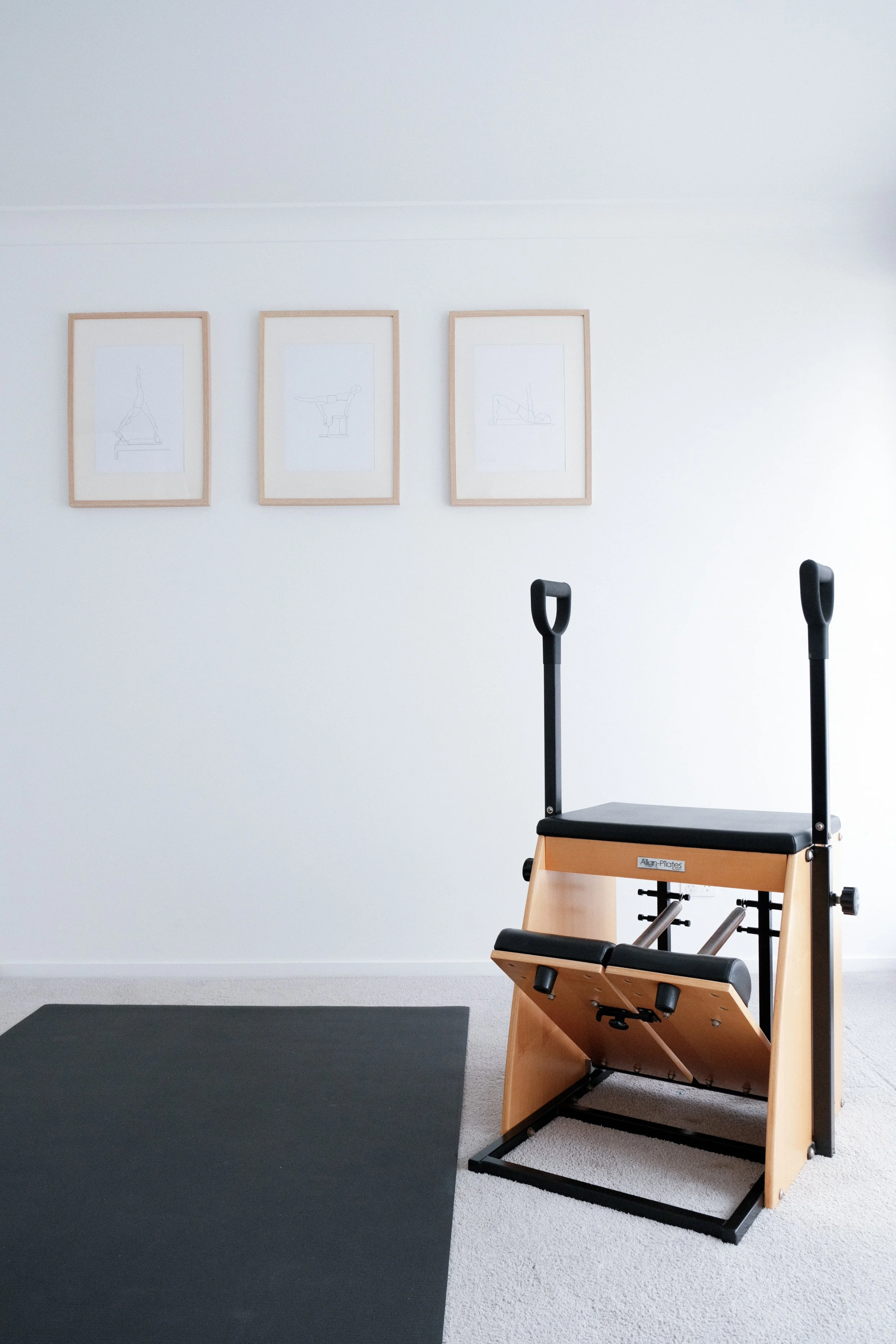 A Pilates reformer machine in a minimalist room with three framed line art sketches on the white wall behind it and a black exercise mat on the light-colored carpeted floor.