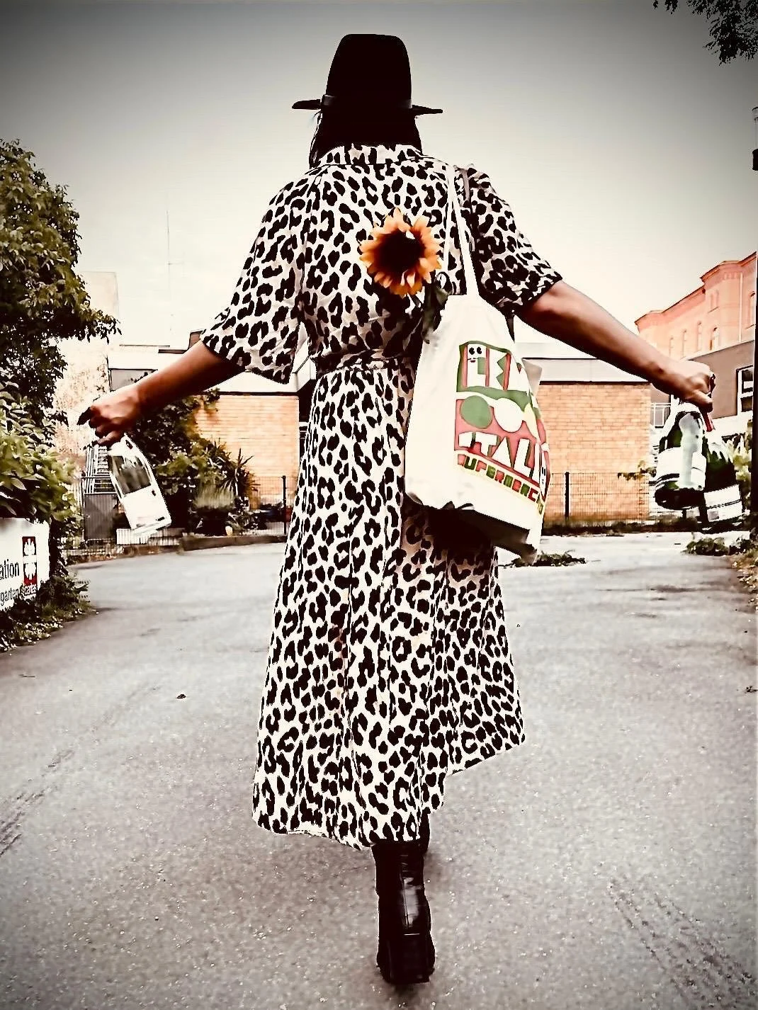 Eine Person trägt ein Leoparden-Muster-Kleid, einen Hut, trägt eine Sonnenblume an der Tasche und hält zwei Flaschen in den Händen, geht auf einer Straße.