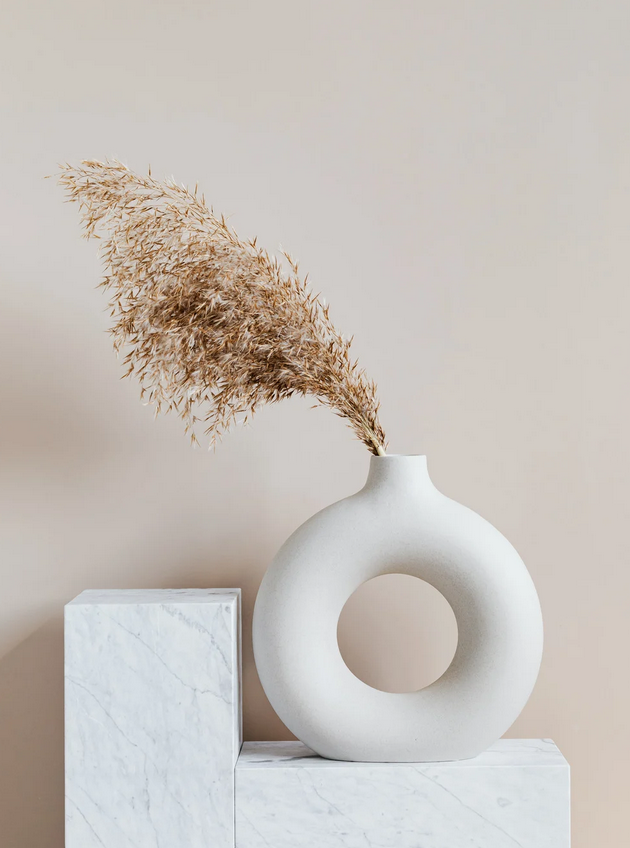 Weiße, runde Vase mit großem Loch in der Mitte, enthält eine getrocknete Pampasgras. Vase steht auf einem weißen Marmortisch gegen eine hellbeige Wand.