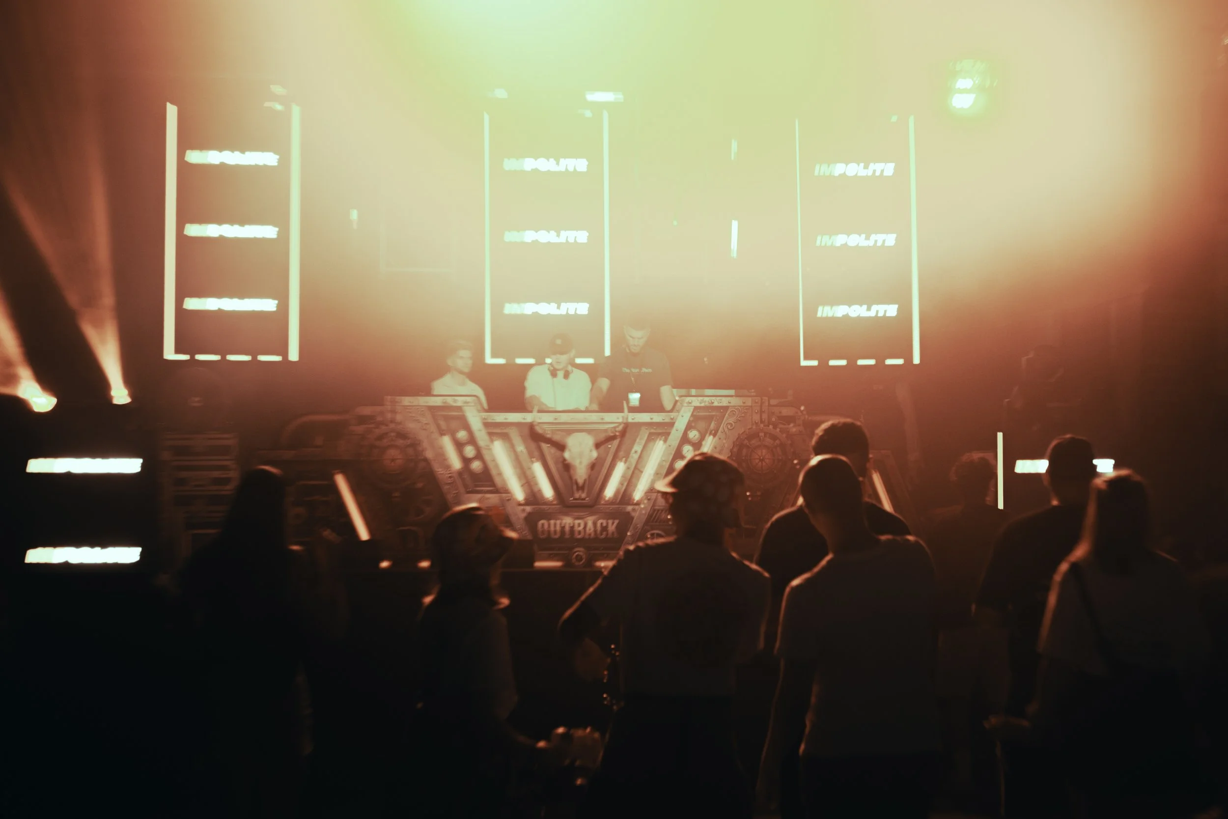 DJ-Team bei einem Musikauftritt vor einer tanzenden Menschenmenge, umgeben von orangefarbenen Bühnenlichtern und großen LED-Bildschirmen.