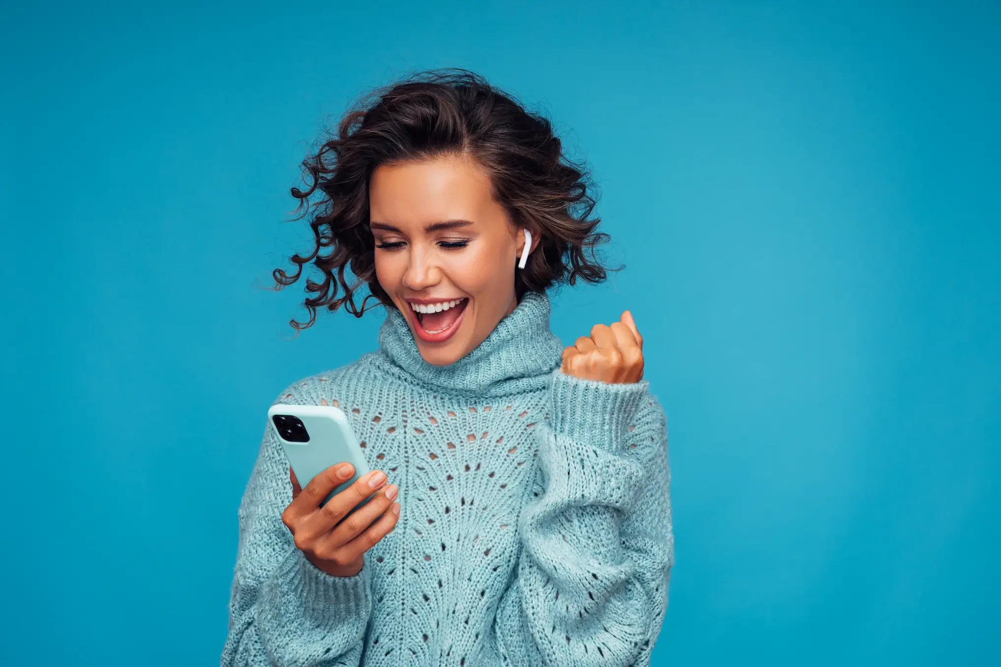 Happy woman with short curly hair celebrating while looking at her phone, wearing a chunky knit sweater and wireless earbuds, against a solid blue background.