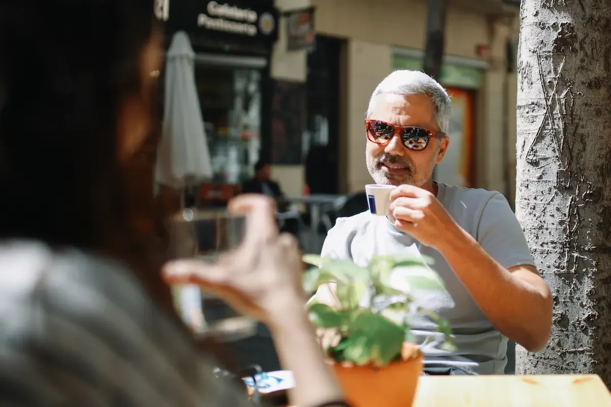 An older man with sunglasses enjoying a cup of coffee at an outdoor cafe, smiling at a woman whose hand is visible holding a similar cup, with a blurred background of buildings and a tree.