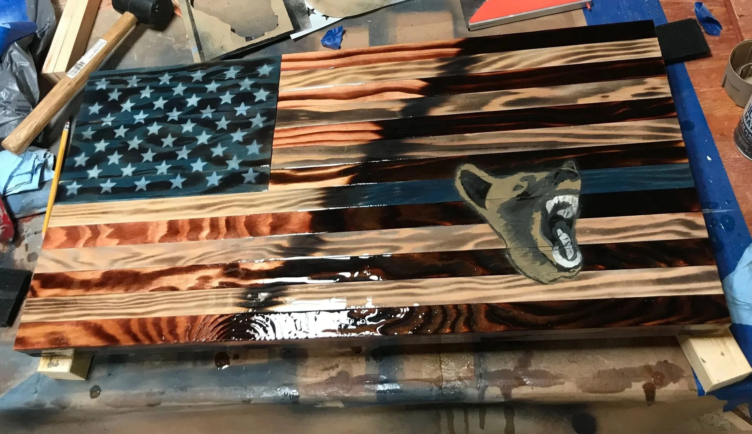 A wooden American flag with burned and stained stripes and a blue canton with white stars, featuring a painted roaring bear face on the lower right side.