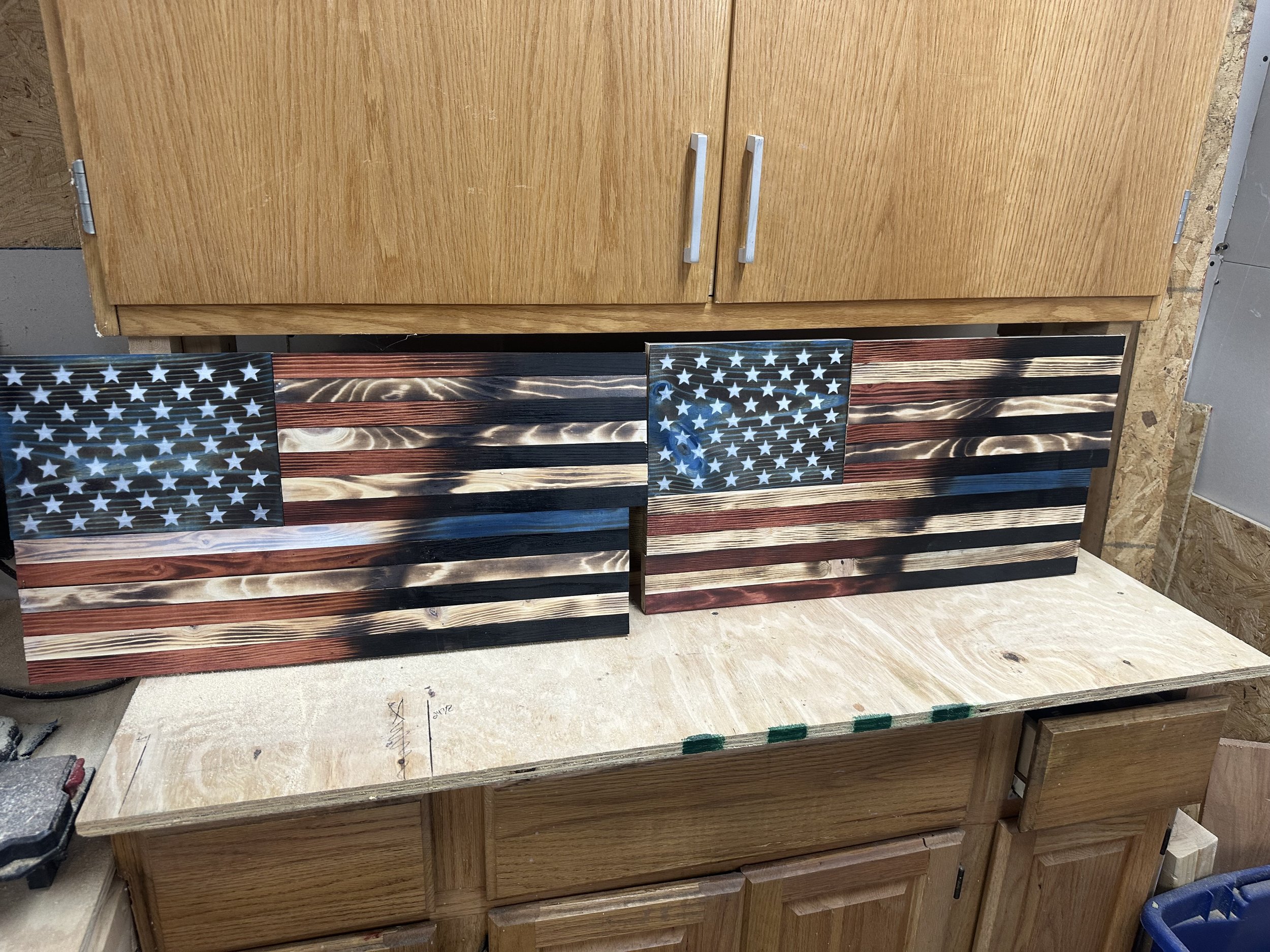 Two wooden American flags with star-spangled blue squares and red and white stripes, propped against a wooden cabinet on a workbench.
