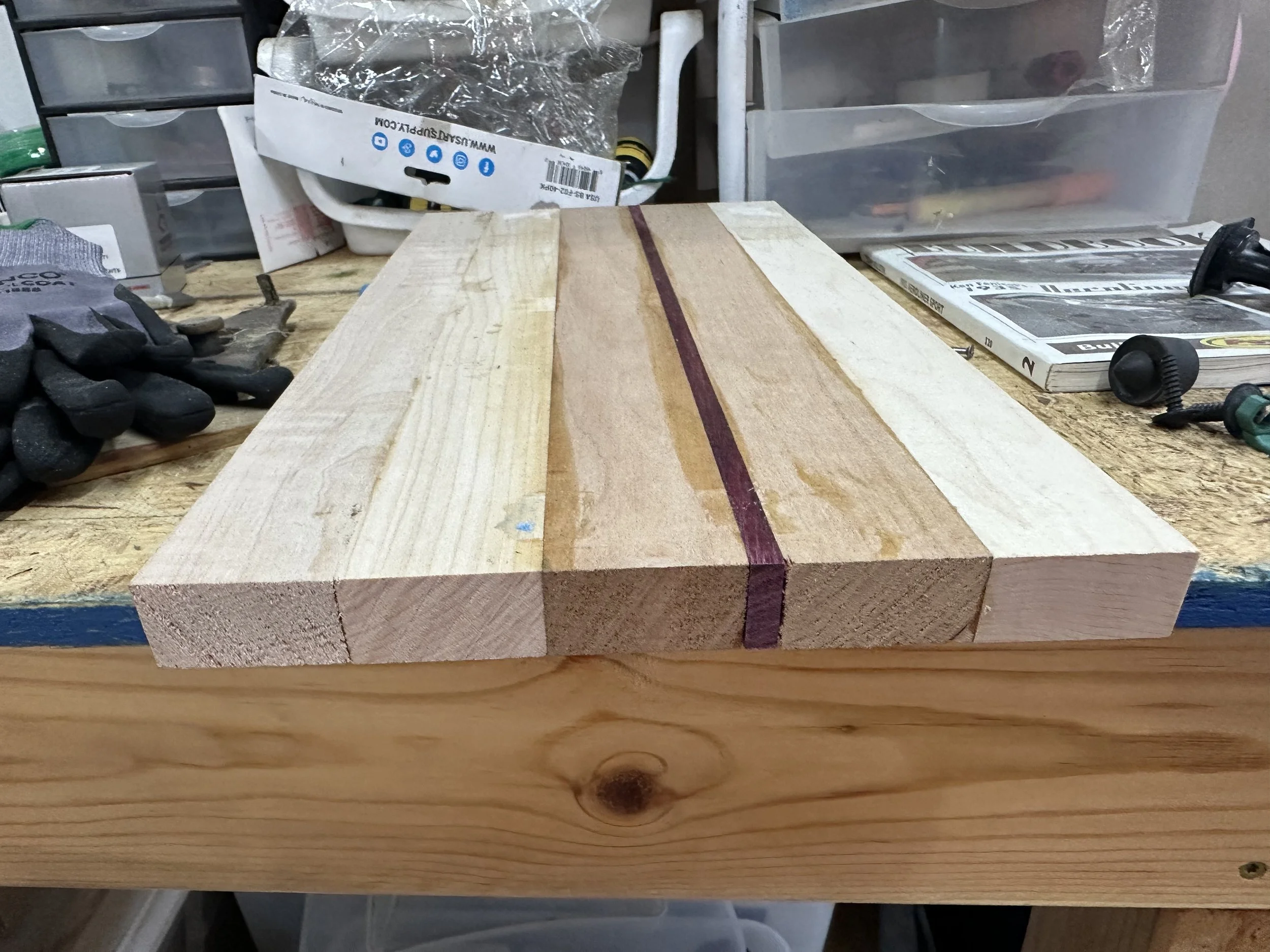 A workbench with a partially assembled wooden project, featuring three pieces of wood joined together with one darker strip in the center, surrounded by tools and materials in a workshop setting.